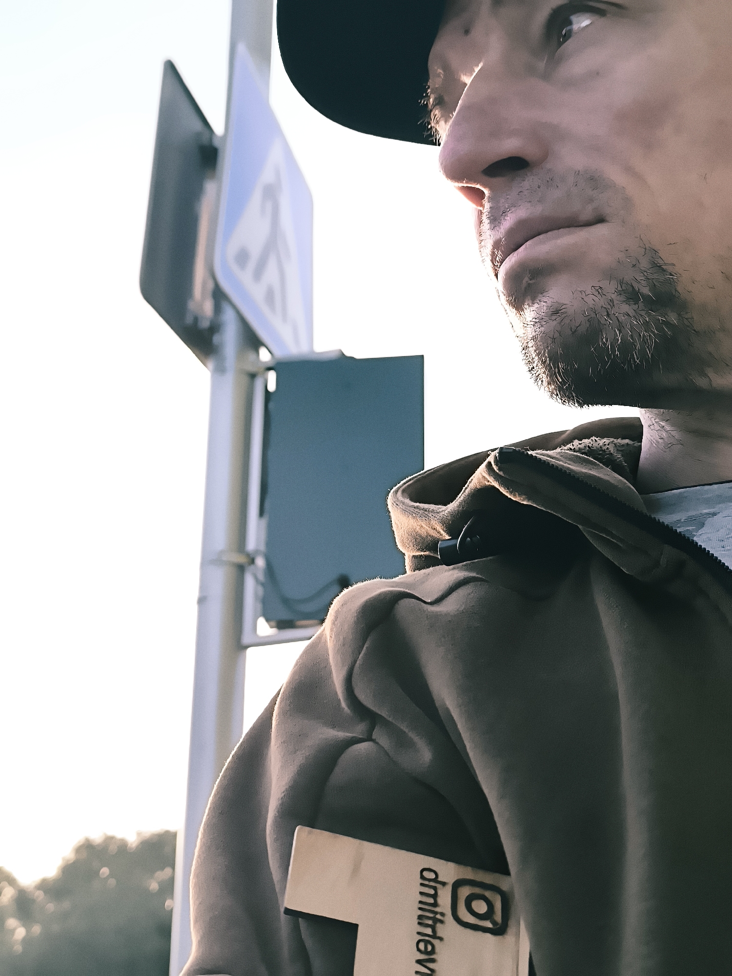 A close-up, profile shot of a man wearing a black baseball cap and a brown hooded jacket, stands in front of a pedestrian crossing sign in Qarağandı city administration, Kazakhstan. The man's face is partially illuminated by sunlight, casting highlights on his nose, lips, and chin, which are covered with stubble. He is looking off to the right, with his mouth slightly open. His right arm is bent, and a small, light-colored rectangular object with black text and an Instagram logo is visible tucked into the front of his jacket. Behind him, the blue pedestrian crossing sign is visible, mounted on a metal pole. The background is largely taken up by a bright, overcast sky and the soft focus of distant trees.