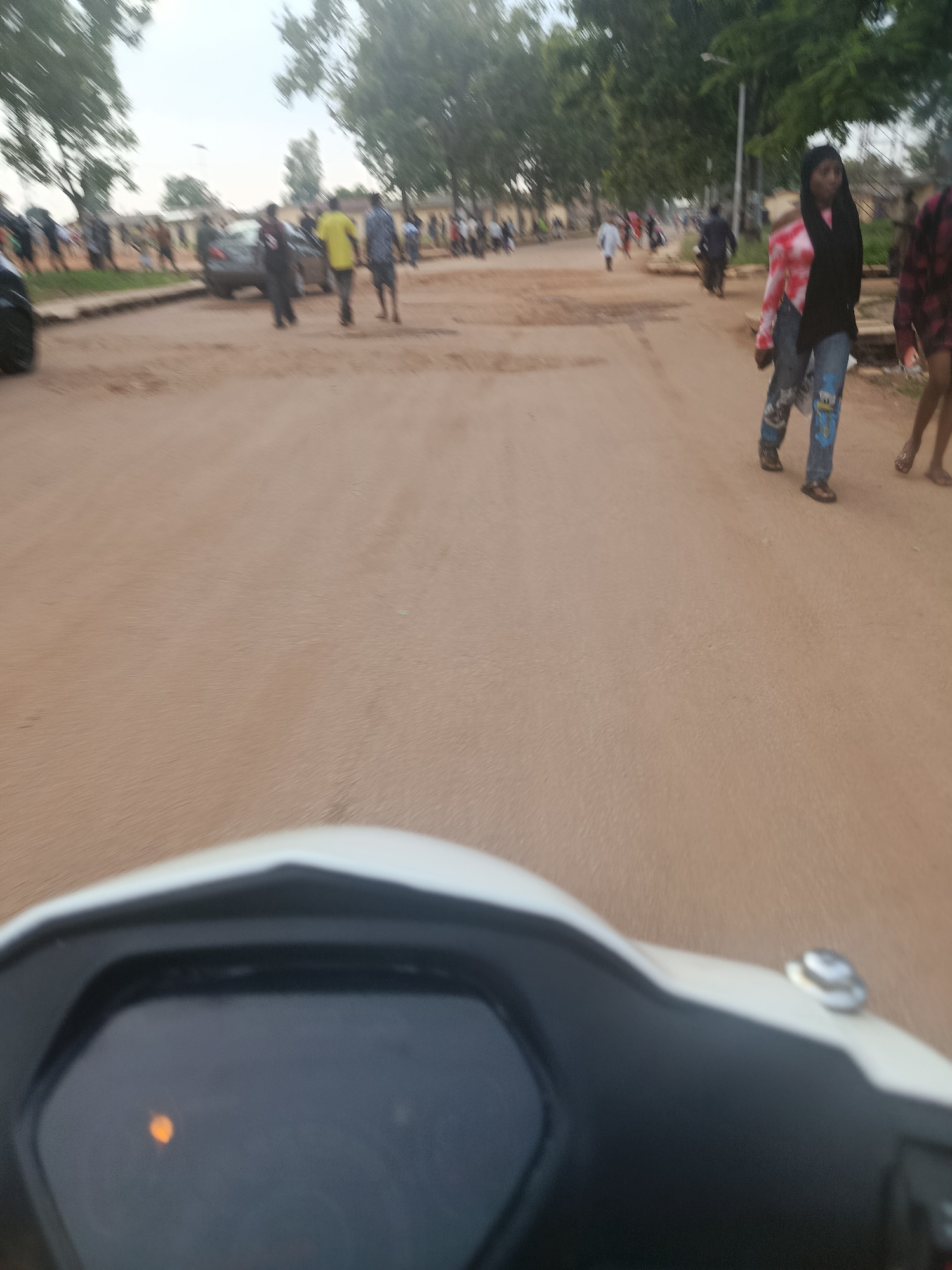 The image captures a street scene in New Stadium, Nigeria, with a focus on a dirt road and a procession of people. A white motorcycle dashboard is visible in the foreground, with a small orange light illuminated. Beyond the dashboard, the dirt road stretches into the distance, lined by trees and buildings. Several individuals are walking on the road, some appearing to be in groups, while others are walking individually. They are dressed in casual clothing. A dark-colored car is parked on the side of the road. The overall atmosphere appears to be a typical day in a Nigerian city, with a sense of movement and activity.