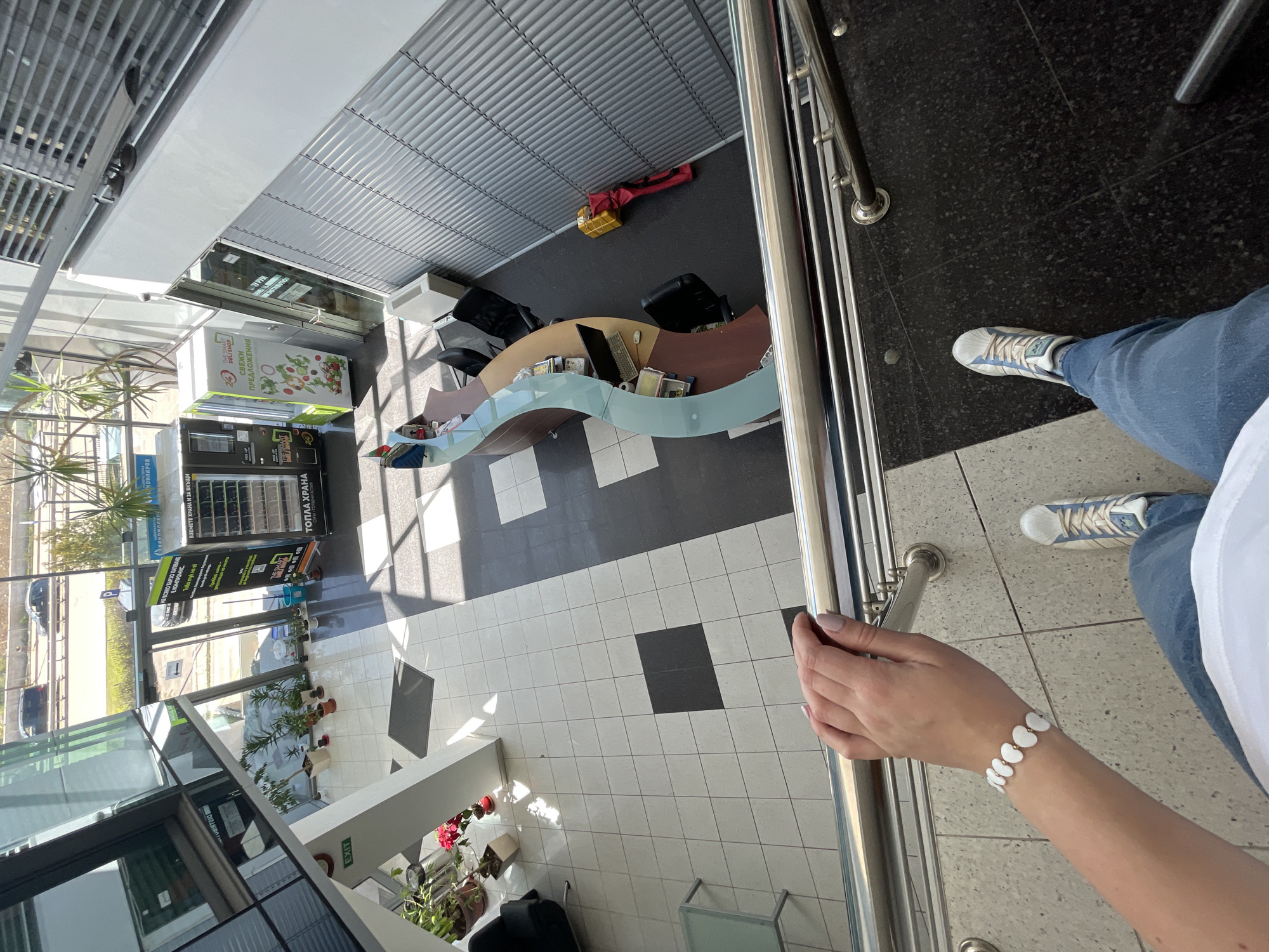 Here's a detailed description of the image:
**Scene Overview:**
The photograph is taken from an elevated perspective, looking down into what appears to be the interior of a commercial building or office space. The angle suggests the photographer is likely positioned on an upper floor or mezzanine.
**People:**
The image includes a portion of a person, specifically an arm and part of their leg. The person is wearing blue jeans, white and blue sneakers, and a white shirt or top. They also have a white bracelet, possibly with heart-shaped beads. The person's hand is holding onto a metal railing.
**Objects:**
* **Furniture:** There is a curved reception desk or counter visible, made with a wood-like material and possibly glass panels. There are black office chairs near the desk.
* **Building Structure:** The interior features a combination of tiled flooring (black and white), metal railings, and large windows, allowing for ample natural light. A metal roof structure with horizontal ventilation grates is also visible.
* **Retail Elements:** A "Deli Shop" stand, vending machines labeled "TOPLA HRANA" (presumably meaning "Hot Food"), and plants/flowers are visible. There is a sign written in Cyrillic that indicates "TAKE FOOD AND FOR HOME".
* **Miscellaneous Items:** There are red and yellow bags laying around.
**Actions and Interactions:**
The person is holding onto the railing, suggesting they are observing the scene below or navigating the space.
**Environment:**
* **Lighting:** The interior is brightly lit, mostly from natural light coming through the windows.
* **Layout:** The building interior has an open layout with a reception/service area and some retail components.
* **Exterior View:** Through the windows, one can see a street with passing cars and possibly greenery or trees outside.
**Sofia, Bulgaria Location Inference:**
The location is likely in Sofia, Bulgaria, because the signs "TAKE FOOD AND FOR HOME" and "TOPLA HRANA" are written in Cyrillic. Cyrillic is the official script of Bulgaria.