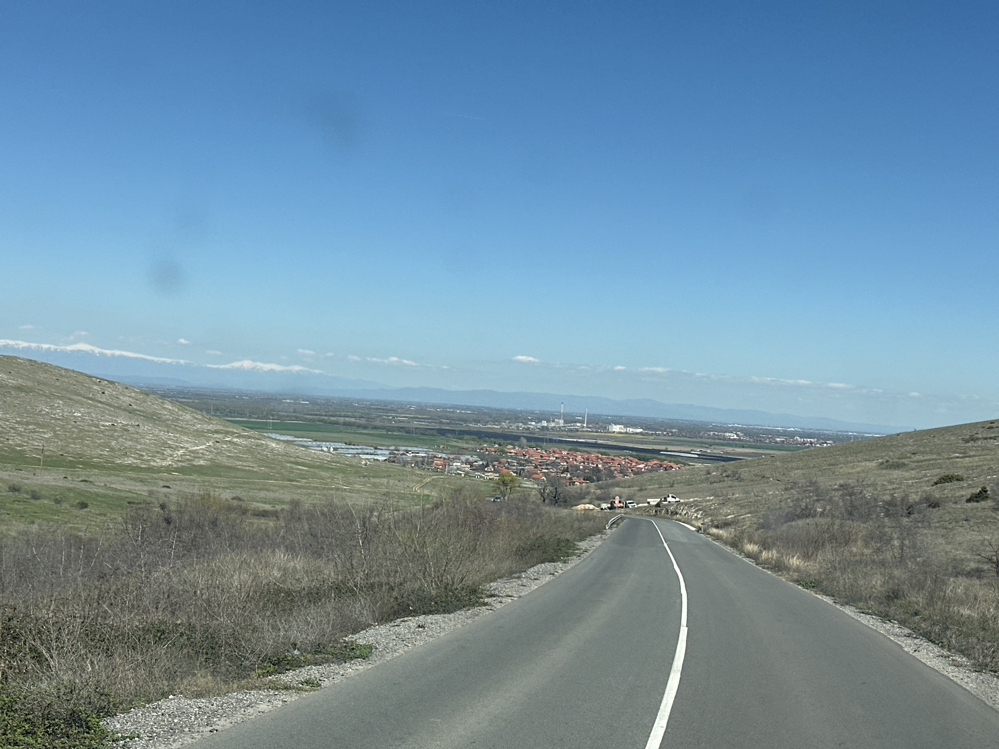 The image captures a paved road with a solid white line marking the lanes, leading towards a small town in the distance. The location is in the city of Isperihovo, Bulgaria. The road is flanked by grassy hills on either side, with dry vegetation close to the roadside. In the background, there's a broader landscape featuring cultivated fields, buildings, and industrial structures. Mountains with snow-capped peaks are visible on the horizon. The sky is clear and blue.