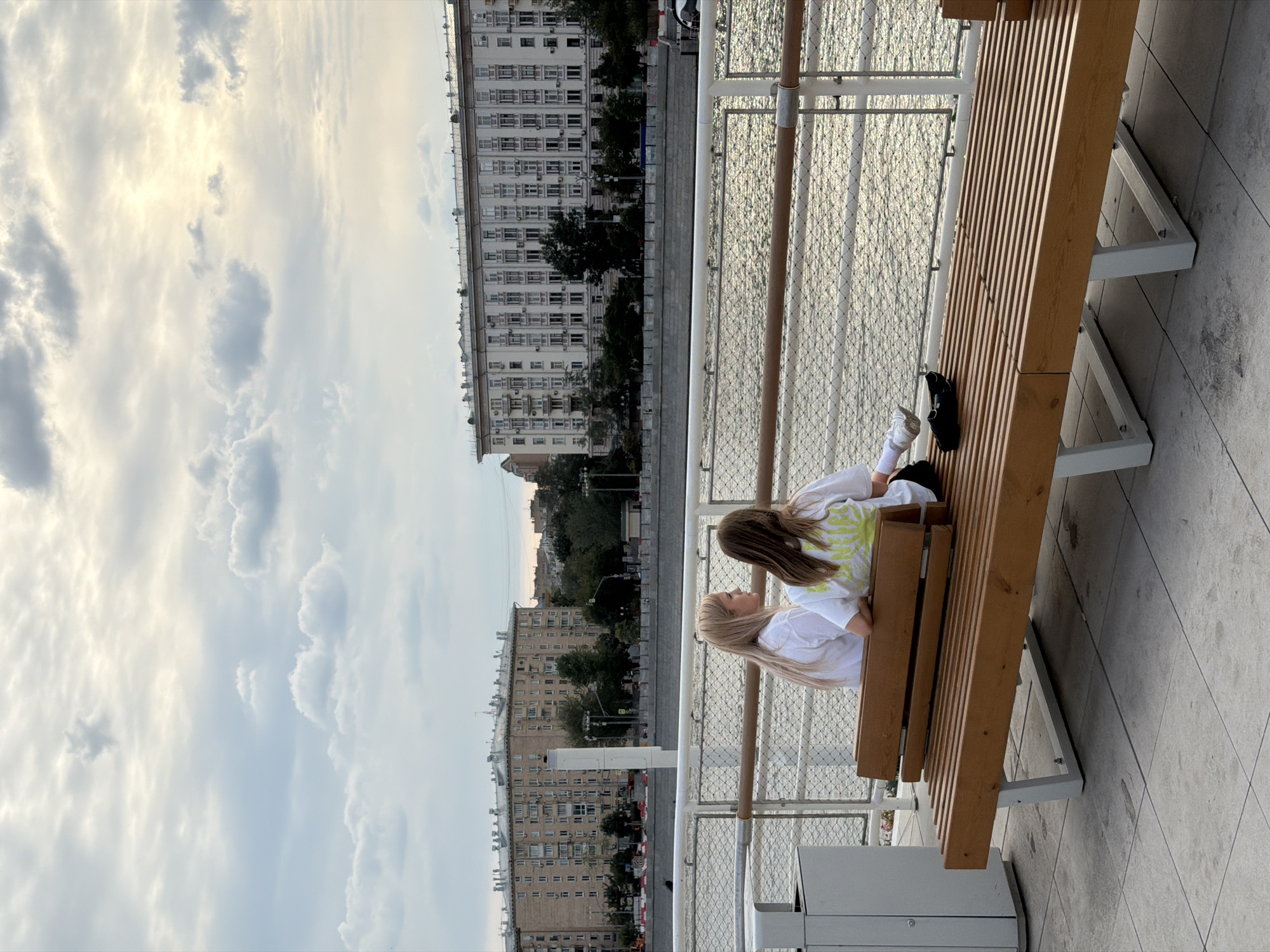 This image shows two young girls sitting on a wooden bench next to a body of water. The girl closer to the camera has long blonde hair and is wearing a white t-shirt. The girl further away has long brown hair and is wearing a white t-shirt with a yellow tie-dye pattern. They are both facing away from the camera, looking out at the water and the city skyline.
The scene appears to be a promenade or waterfront area, with a railing separating the bench from the water. In the background, there are several large, multi-story buildings, characteristic of urban architecture. The sky is cloudy, with patches of sunlight breaking through, suggesting it might be late afternoon or early evening. The water's surface is reflecting the light from the sky.
The overall mood seems relaxed and contemplative, with the girls enjoying the view. There is no visible text in the image. Based on the architectural style of the buildings and the presence of a river, the location is likely Moscow, Russia, as indicated by the location context. The presence of the railing and the modern bench suggests a public recreational area.