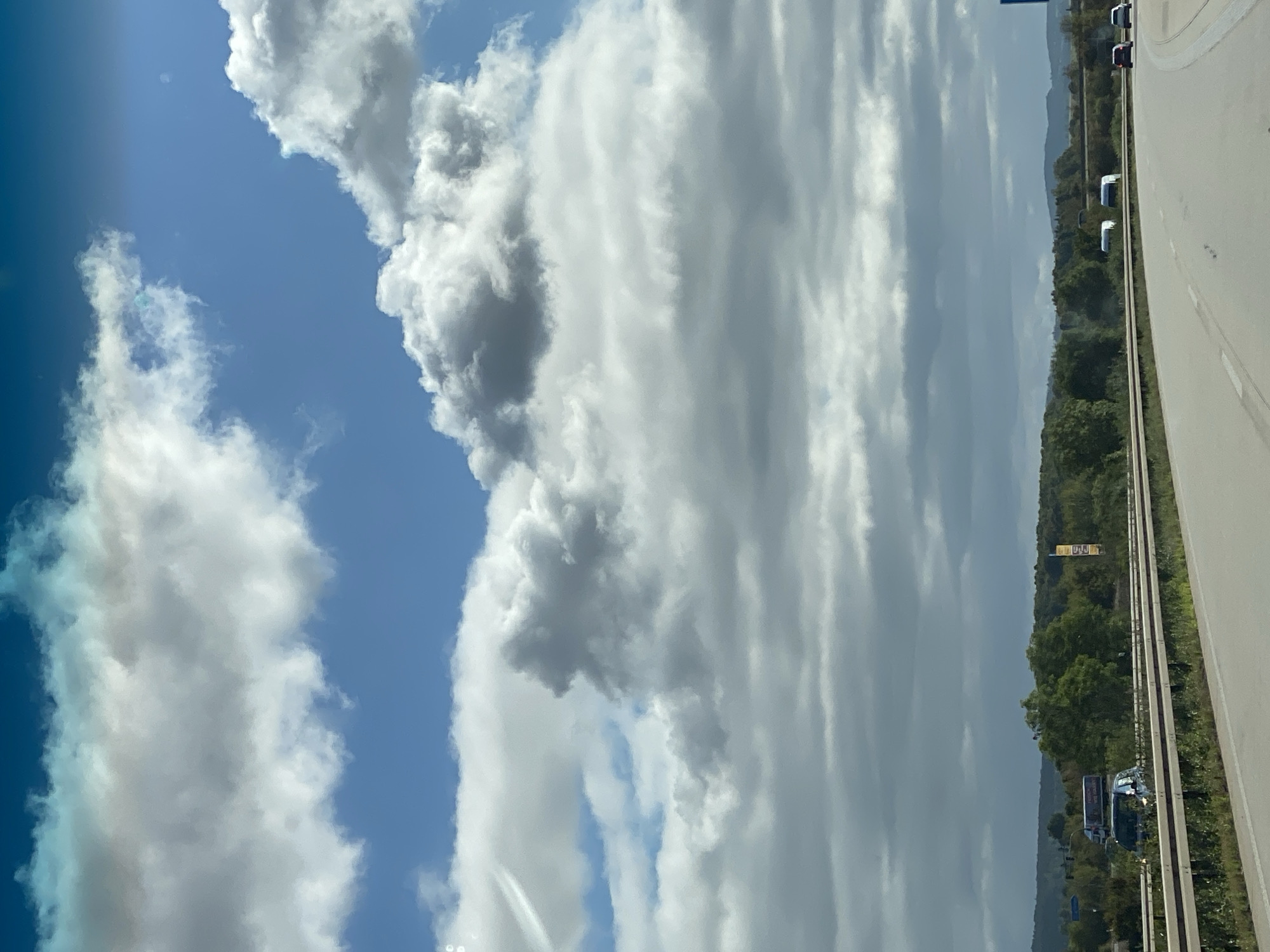This is a photograph taken from a vehicle traveling on a highway. The sky is filled with a mix of fluffy white and darker gray clouds, with patches of blue sky visible. Below the sky is a landscape with trees and hills. A guardrail runs along the side of the highway, and several vehicles, including cars and what appear to be trucks or vans, are visible on the road. The overall scene suggests an overcast day with some sun peeking through. The location is the A38 highway near Heringen/Helme, Germany.