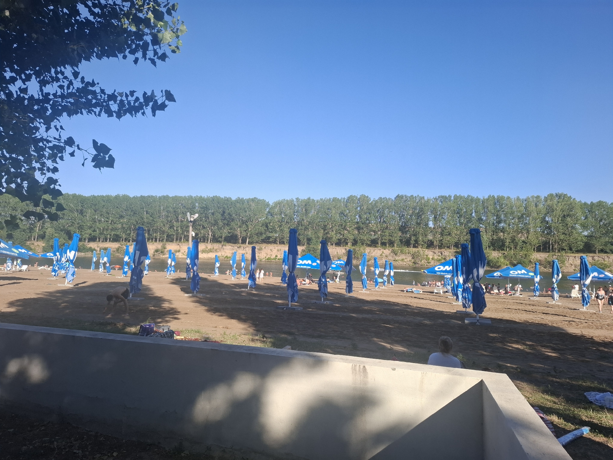 The image depicts a crowded beach scene in Vadul lui Voda, Moldova, on a clear, sunny day. The foreground is dominated by a concrete structure casting shadows, with some foliage on the left. Beyond this, a sandy beach stretches towards a river, lined with numerous closed blue umbrellas. Several people are visible on the beach and in the water, some sunbathing on lounge chairs and others swimming. In the middle ground, a young person is bending down on the sand, possibly playing or interacting with something on the ground. The background features a dense line of trees along the riverbank. The overall atmosphere is that of a recreational area, likely a popular summer destination.