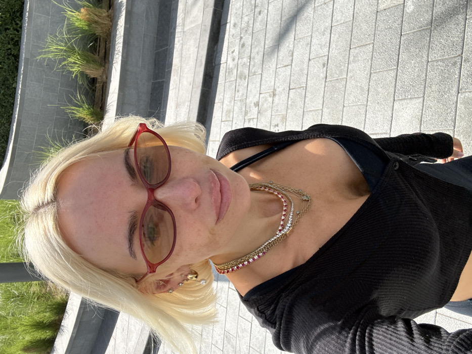 A fair-skinned female with shoulder-length blonde hair, parted centrally, is depicted in a close-up outdoor selfie. She is wearing small, oval-shaped red-framed sunglasses with tinted lenses. Her face exhibits some freckles across the nose and cheeks. Her ears are adorned with multiple small earrings, including a gold hoop and a dangling pearl-like earring on her left ear. Around her neck, she wears several layered necklaces, including a prominent one with red and white stones, and several gold-toned chains, one featuring a small green charm. She is dressed in a black, long-sleeved ribbed top with a V-neckline, worn over a dark undershirt or bra, revealing some cleavage. The lighting indicates bright daylight, casting distinct shadows. The background consists of a paved walkway made of light grey rectangular blocks. Beyond the walkway, there is a terraced planter area with curved grey stone walls, featuring clusters of green ornamental grasses. A dark, vertical pole is visible in the upper left background. The scene is located in Dubai, United Arab Emirates.