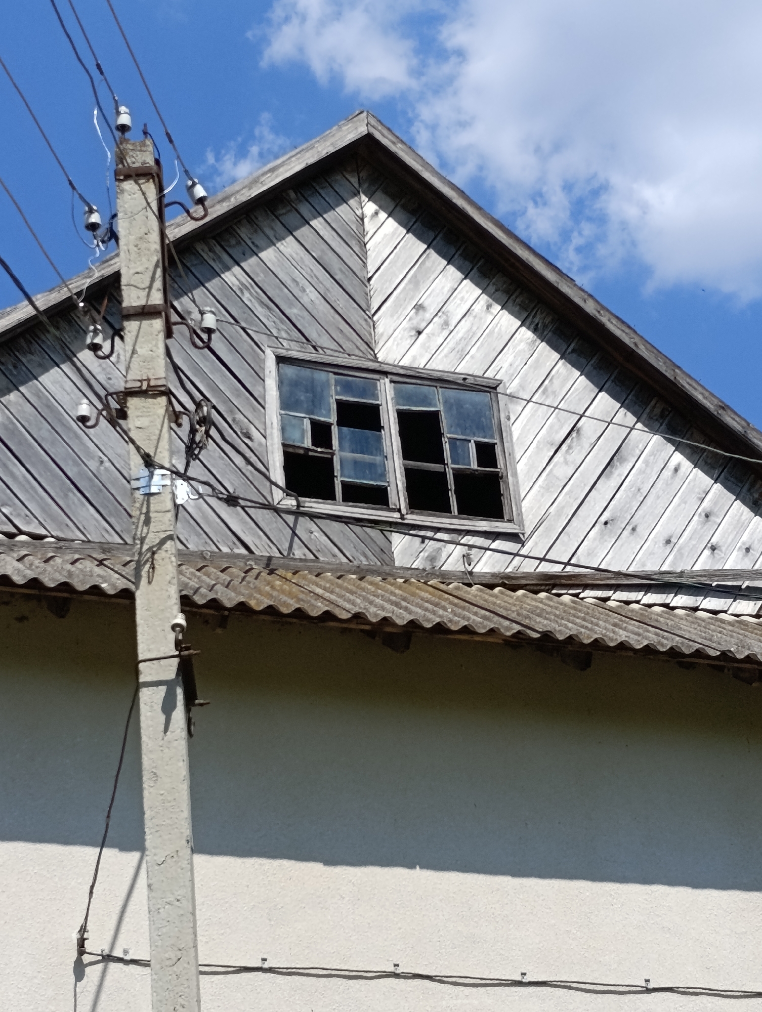 The image shows a close-up of an abandoned wooden house in Koikovo, Moldova, under a bright blue sky with scattered clouds. A concrete utility pole with overhead wires and ceramic insulators stands in the foreground to the left of the house. The house has a steep, weathered wooden roof with diagonal siding. A single, broken window is visible in the gable end of the roof, with multiple panes missing or shattered. The corrugated roof of a lower section of the house is visible below the gable. The overall scene suggests decay and neglect.