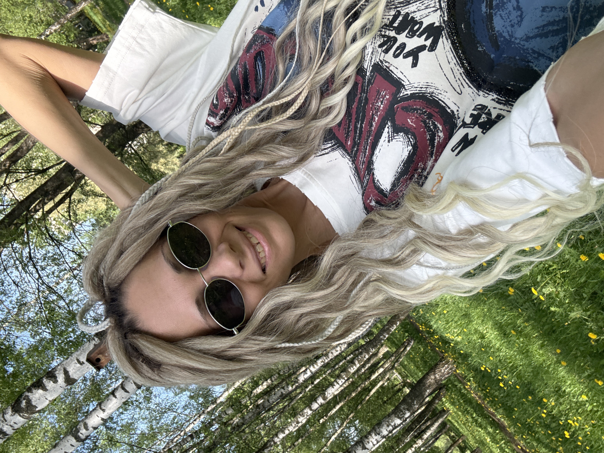 A fair-skinned woman with blonde braids and sunglasses lies on her back in a grassy field dotted with yellow flowers. The woman is wearing a white t-shirt with a graphic print. Above her, a forest of birch trees stretches towards a blue sky. The location is presumed to be in a park or green space in Rusanovo, Russia, based on the prompt.