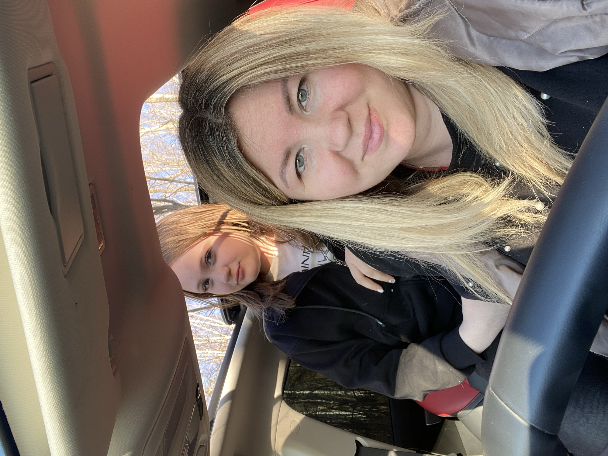 This brightly lit selfie, taken from a slightly tilted perspective inside a car, features two females of different ages, likely a mother and daughter. The elder woman, positioned in the foreground, is holding the camera. She has long, straight blonde hair with visible darker roots and striking green eyes, and she offers a gentle, subtle smile to the lens. She is wearing a dark top, possibly adorned with pearl-like details visible near her neckline.
In the background, the younger female is seated further back in the passenger area, looking towards the camera with a neutral, slightly contemplative expression. She has lighter brown hair. She wears a dark hooded jacket over a light-colored t-shirt, on which the partial text "ONI NT" is visible. A red bag or object is partially seen near her lap.
The setting is the interior of a vehicle, with parts of the car's roof lining and a black steering wheel visible. Abundant natural light enters through the windows, illuminating the subjects and the cabin. Through the windows, a clear blue sky and the silhouettes of numerous bare tree branches are discernible, indicative of either late winter or early spring weather in Chaikovsky, Russia. The image captures a casual, everyday moment between the two individuals.