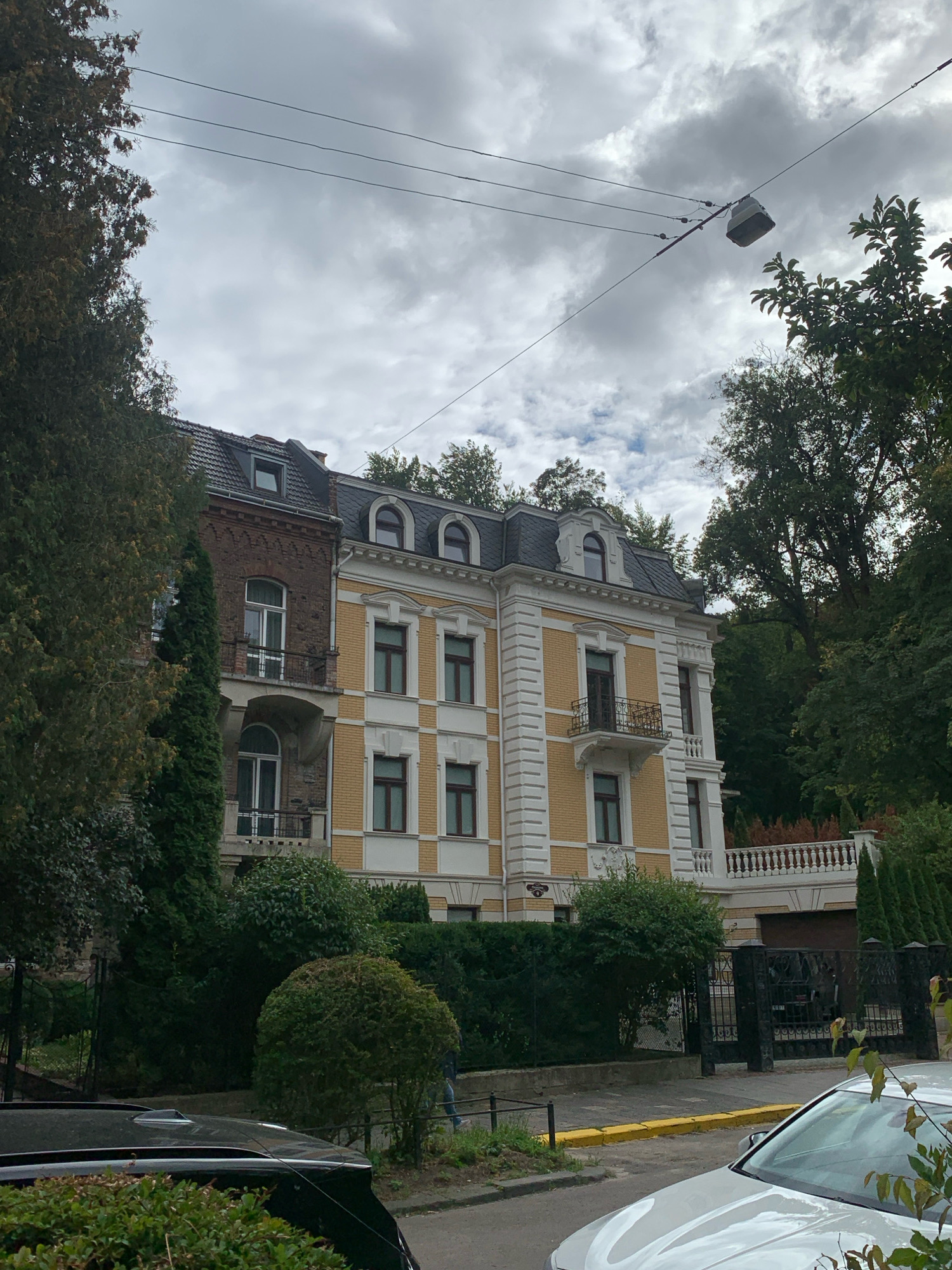 The image shows a multi-story building with a yellow facade and ornate white trim, situated in Lviv, Ukraine. The building has arched windows and balconies with decorative railings. Adjacent to it, a portion of a darker brick building is visible, also with arched windows. Both structures appear to be residential or commercial properties with a classic architectural style.
The building is surrounded by lush greenery, including mature trees and well-maintained hedges. A black wrought-iron fence runs along the property line, with a large gate in the foreground. There are at least two cars visible, one black and partially obscured by foliage in the lower left, and another white car in the lower right foreground, positioned on a paved street. A yellow curb separates the street from the sidewalk. Overhead, power lines crisscross the sky, with a street lamp visible. The sky is overcast with dramatic, cloudy formations. The overall impression is that of an affluent urban neighborhood with substantial, well-kept properties.