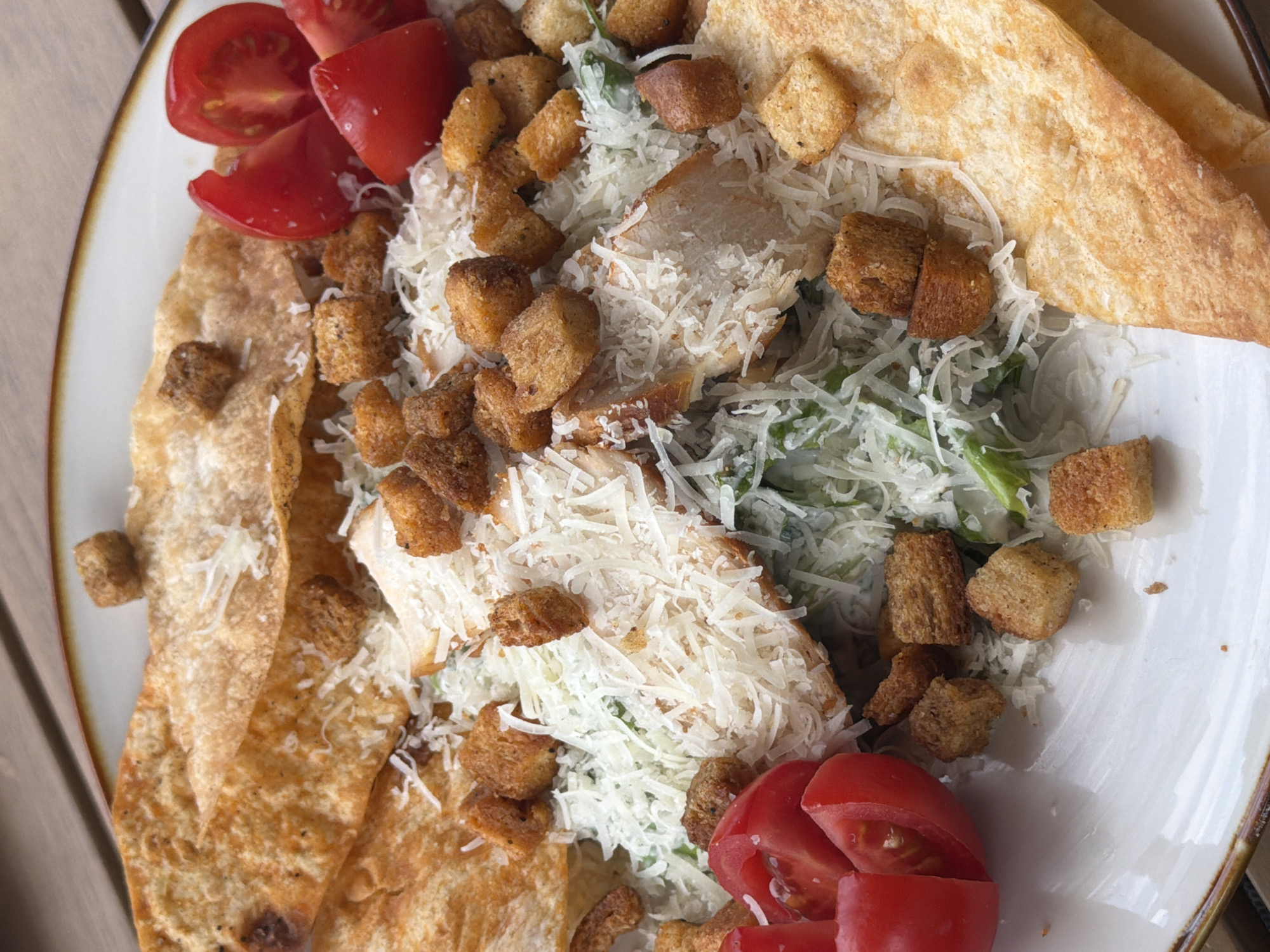 This is a close-up, overhead shot of a Caesar salad on a white plate. The salad features grilled chicken strips, croutons, shredded Parmesan cheese, and sliced cherry tomatoes. Large, golden-brown tortilla chips are layered around the salad. The plate is set on a wooden surface, likely a table. The background is slightly blurred, suggesting an outdoor or well-lit indoor setting. There are no people visible in the image. This scene is located in Byurakan, Armenia.