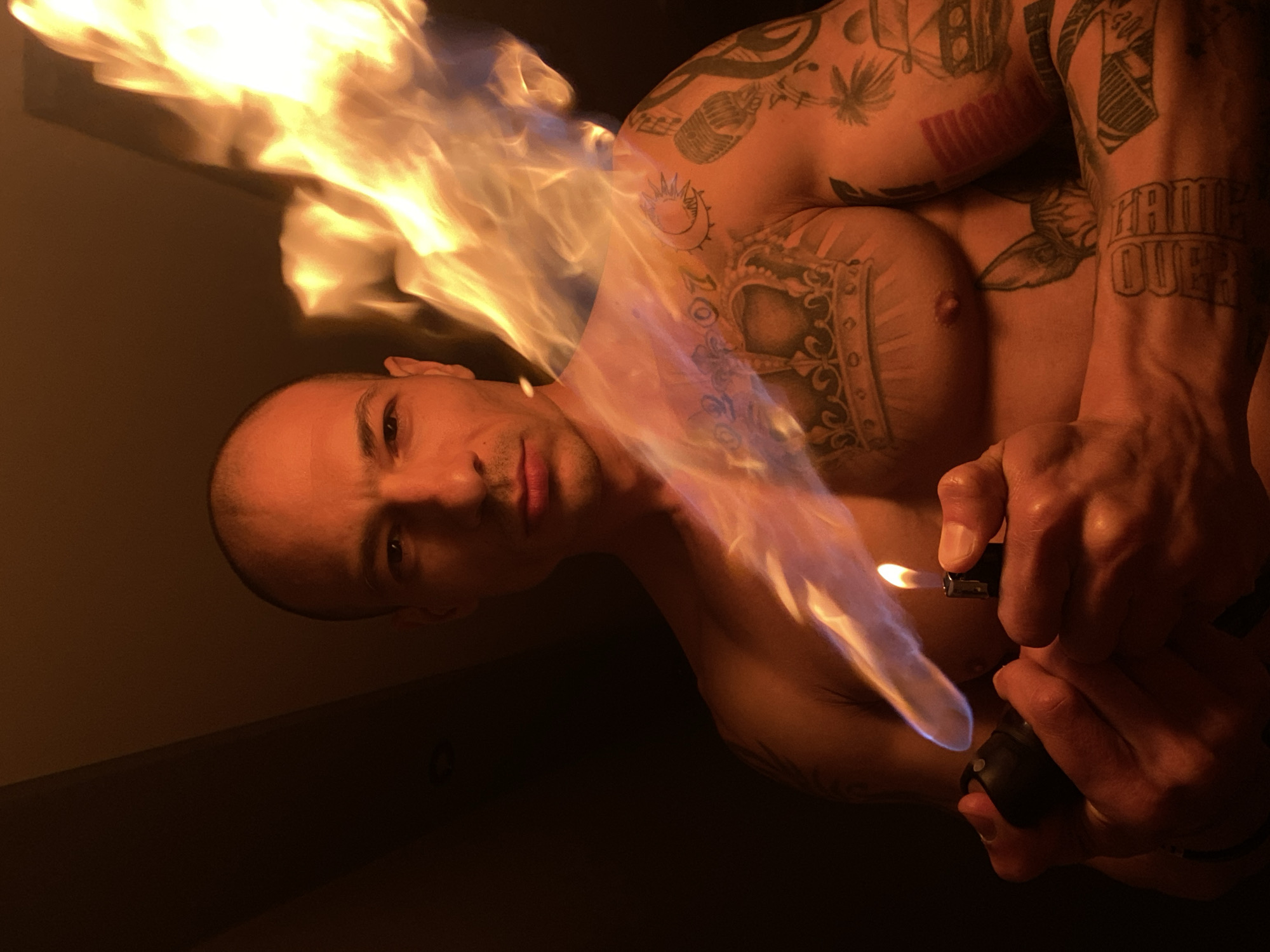 A shirtless man with numerous tattoos is captured in a dark, dimly lit indoor setting in Pleven, Bulgaria. The image is oriented with his head on the left, looking directly at the camera with a neutral and focused expression. A large, bright orange and blue flame erupts from a lighter held in his right hand, positioned near his chest and extending upwards towards his face and the upper part of the frame. The flame acts as the primary light source, casting warm, dramatic highlights on his skin and the surrounding darkness.
The man's body is heavily adorned with tattoos. Prominently visible on his chest is a large, elaborate crown or crest design. His right arm (viewer's left) features a microphone, musical notes, and what appears to be a palm tree design. On his left forearm (viewer's right), the text "GAME OVER" is clearly visible in block letters, surrounded by other geometric and abstract patterns. A sun and moon motif can be seen on his upper left shoulder/back area. The dark background is featureless, emphasizing the man and the intense fire. The activity suggests a controlled performance or stunt involving fire.