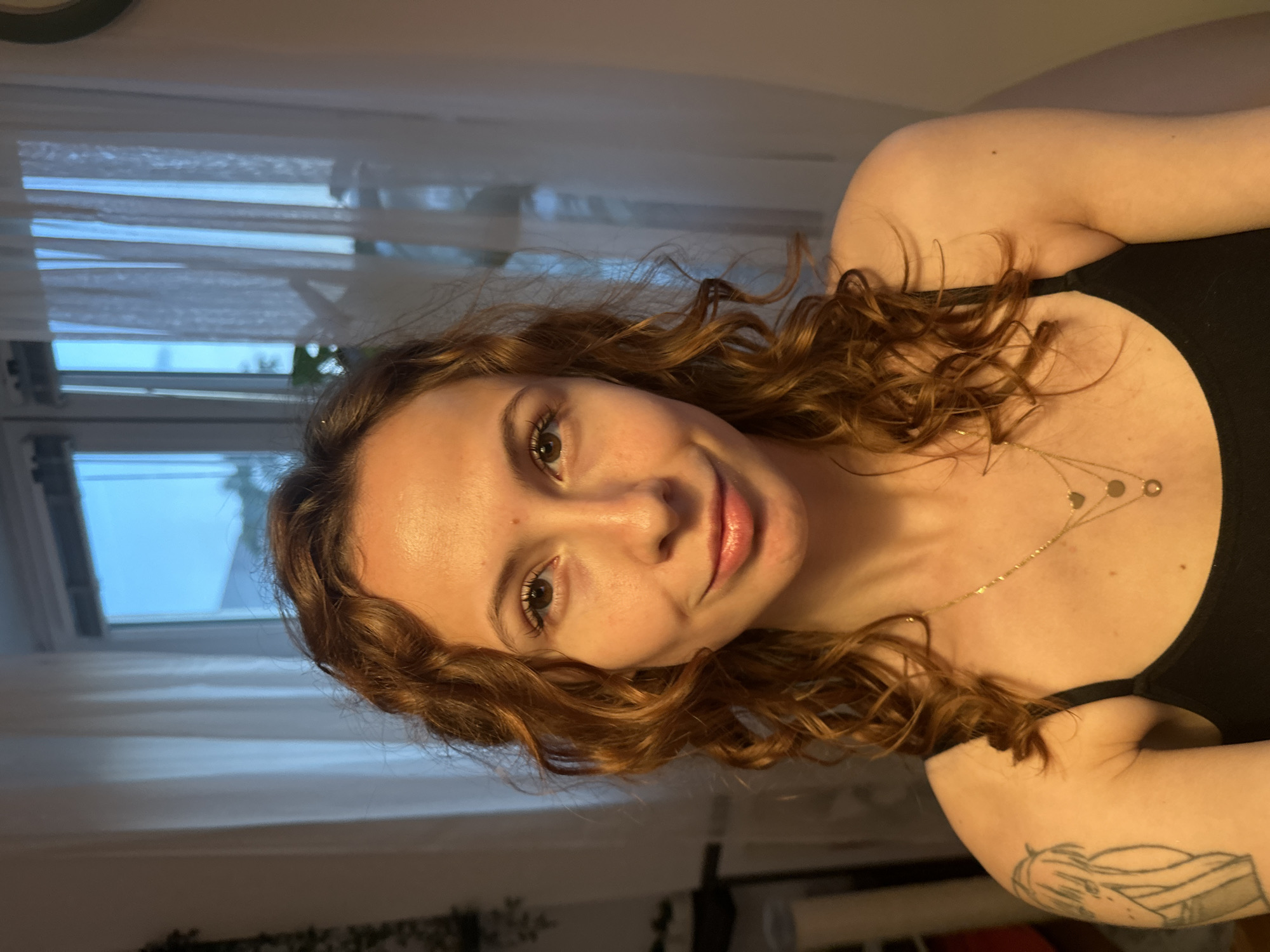 A woman with curly, reddish-brown hair is facing the camera. She is wearing a black tank top and a delicate gold necklace with circular pendants. She has fair skin and light brown eyes. Her lips are slightly parted in a smile. A tattoo is visible on her upper left arm.
Behind her, there is a window with white curtains. The view outside the window is blurry, but it appears to be daytime.
It is impossible to determine the exact location in Novi Sad, Serbia, based on this image alone.