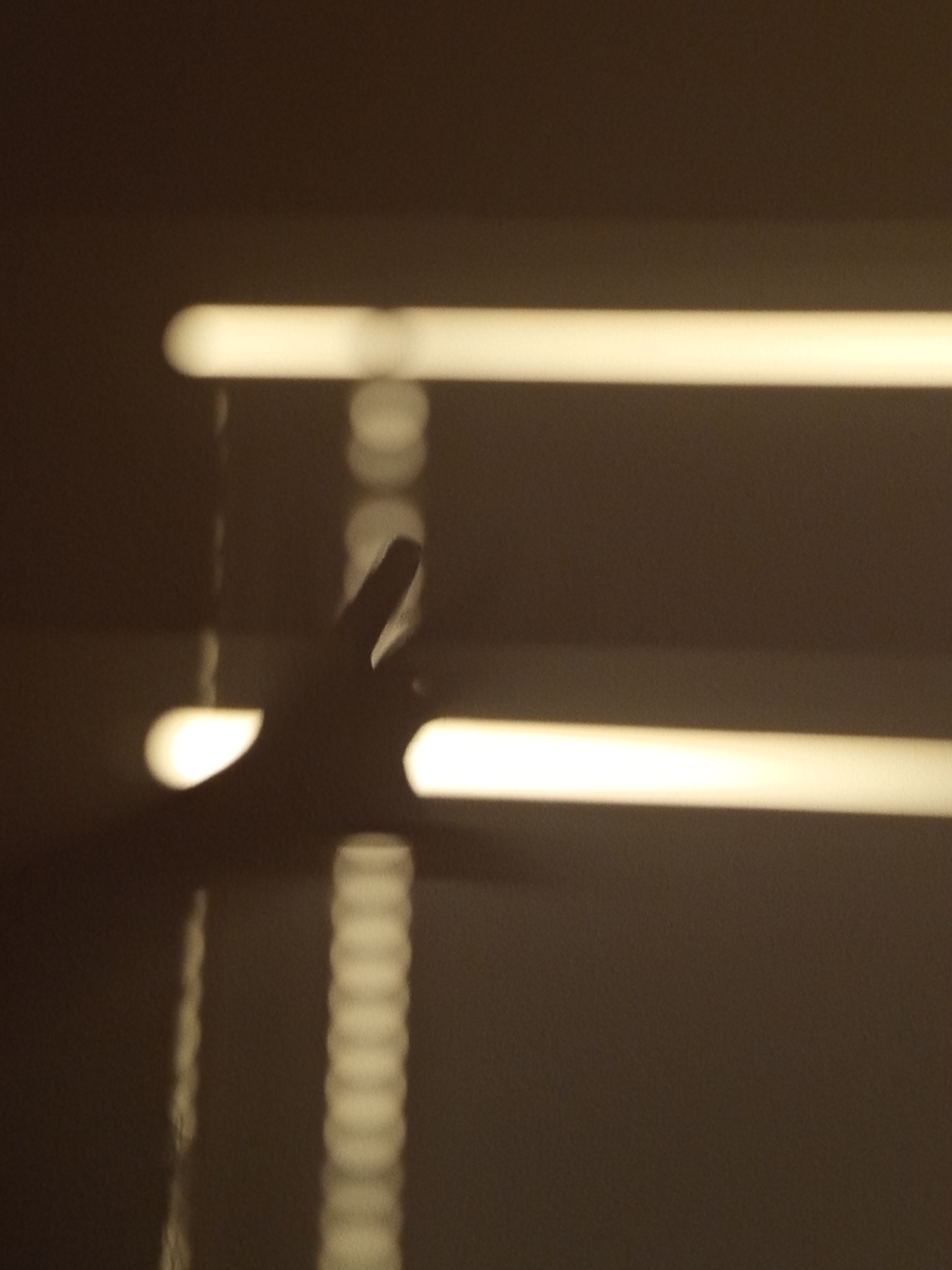 An out-of-focus, dimly lit image shows a hand making a peace sign gesture, silhouetted against a wall. The hand is positioned in the lower-middle of the frame. Two horizontal bands of light, likely from a window or blinds, are visible across the image, casting shadows and creating bokeh effects on the wall. The overall scene suggests an indoor setting at night or in low light conditions. The location is Unknown, Unknown.