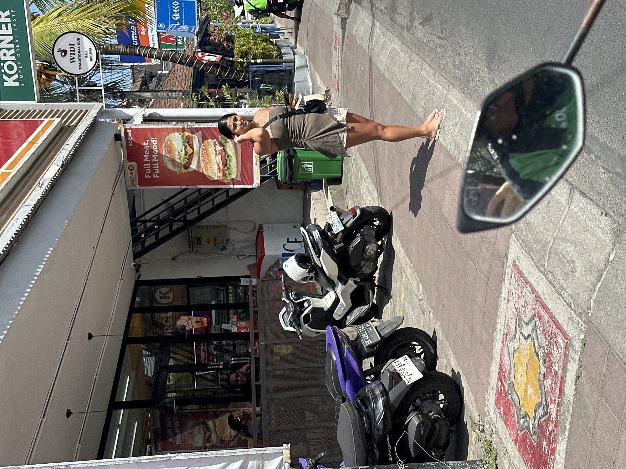A woman stands on a sidewalk in front of a Circle K convenience store in North Kuta, Indonesia. The woman is wearing a brown strapless dress and white shorts. She is holding a phone in her right hand and has her left hand up to her ear. She is barefoot and wearing flip-flops.
In the background, there are several motorcycles parked on the sidewalk. The Circle K store has a large banner advertising burgers. A green trash can is located next to the store. A palm tree is visible to the right of the store. The sun is shining and the scene appears to be during the daytime. The sidewalk is paved with red bricks and there is a grey asphalt road to the right of the sidewalk. There is a man inside the Circle K store visible through the window.