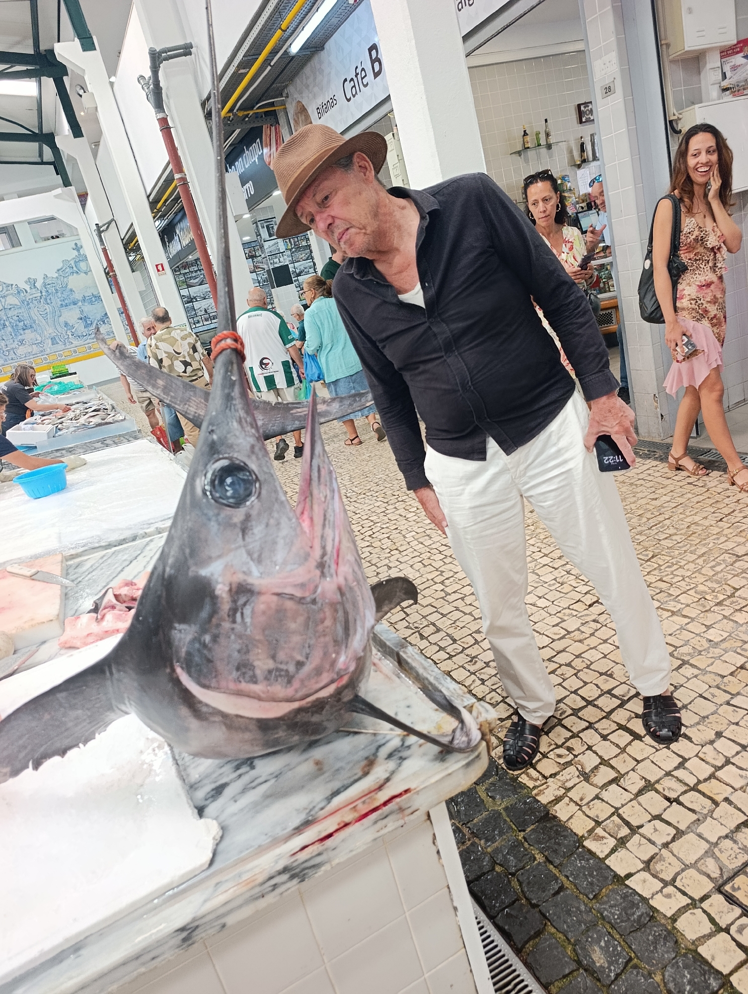 This is a medium shot of a man in a brown hat, a black button-down shirt, and white pants, standing in what appears to be a market in Setúbal, Portugal. The man is looking down at a large swordfish head displayed on a marble counter in the foreground. He is holding a smartphone in his right hand. Behind the man, a woman in a patterned dress is walking away, talking on her phone. In the background, other market-goers can be seen browsing fish and other goods. The market environment includes tiled floors, white pillars, and signage.