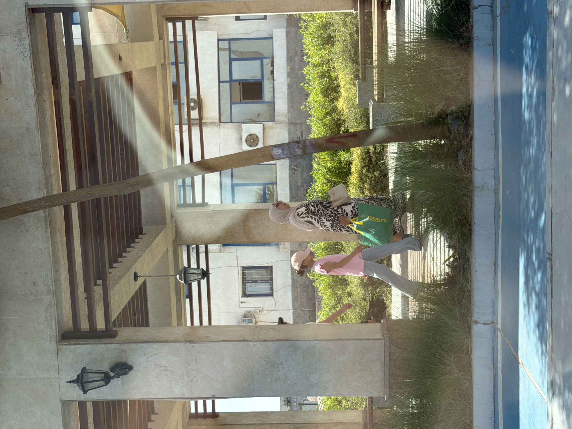 The image captures a street scene in Sharm El Sheikh, Egypt. Two women are walking past a colonnade with a patterned ceiling. The woman on the left wears a hijab and a black and white patterned dress, carrying a cream-colored handbag and a green reusable bag. To her right, a young girl in a pink t-shirt and a pink baseball cap walks alongside her, wearing gray pants. Behind them, a building with multiple windows, some with blue frames, is visible. Lush green bushes line the area in front of the building. The ground in the foreground is a cracked blue surface, possibly a pathway or road, with shadows cast by the overhead structure. A wooden beam stands vertically in the middle of the frame, partially obscuring the scene. The overall lighting suggests a bright, sunny day.