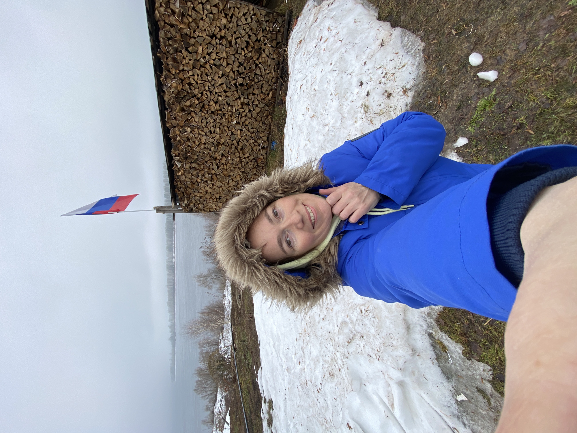 A selfie taken outdoors on a cloudy day, showing a woman wearing a blue coat with a fur-lined hood. Behind her is a stack of firewood, a patch of melting snow, and a view of a body of water. A Russian flag is visible in the background. Based on the location in Ольховка, Russia, it is likely a rural or semi-rural setting near a lake or river.