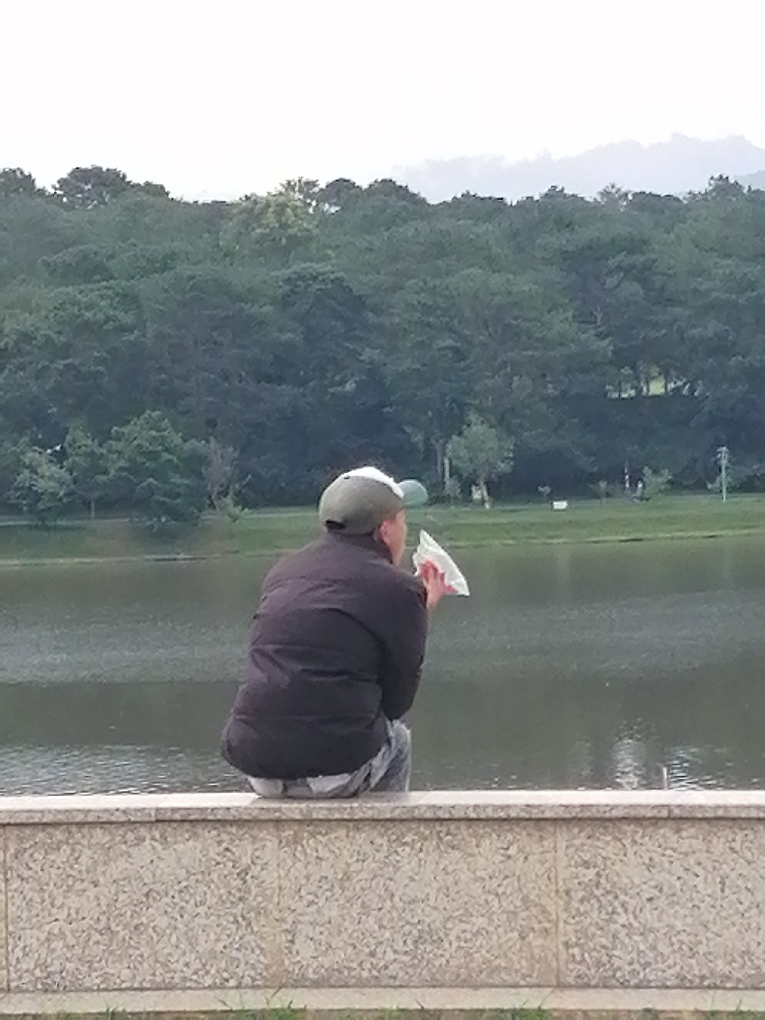A person wearing a dark jacket and a baseball cap sits on a low stone wall overlooking a body of water. The person is holding a white plastic bag up to their face. In the background, a dense line of green trees forms a backdrop against a hazy sky. The water appears still with subtle ripples. The stone wall has a speckled, granite-like texture. The scene suggests a peaceful outdoor setting, possibly a park or lakeside. The attire of the person, the lush vegetation, and the overall atmosphere are consistent with a location in Da Lat, Vietnam, a city known for its natural beauty and cooler climate.