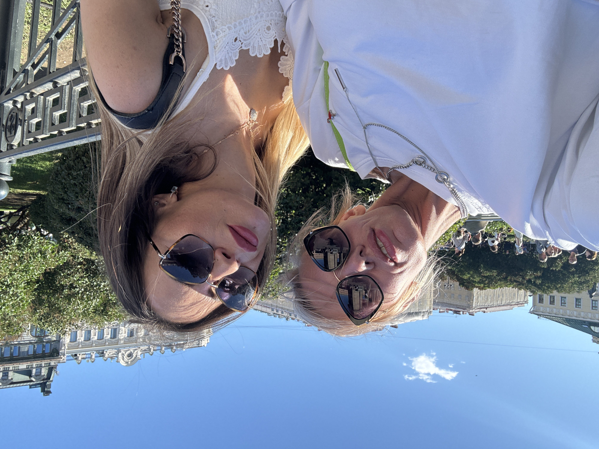 This is a selfie taken by two women outdoors in what appears to be Saint Petersburg, Russia. The photo is rotated 90 degrees clockwise. The women are positioned in the foreground, close to each other, and are looking towards the camera. Both are wearing sunglasses and appear to be smiling. The woman on the left has dark hair and is wearing a white lacy top. The woman on the right has blonde hair and is wearing a white top. Behind them, there is a glimpse of a decorative metal fence, lush green trees, and classical European-style buildings with ornate facades and mansard roofs. The sky is a clear blue with a few wispy clouds, suggesting it is daytime with good weather. In the background, a crowd of people can be seen gathered, possibly for an event or simply enjoying the location. The overall mood of the image is cheerful and relaxed.