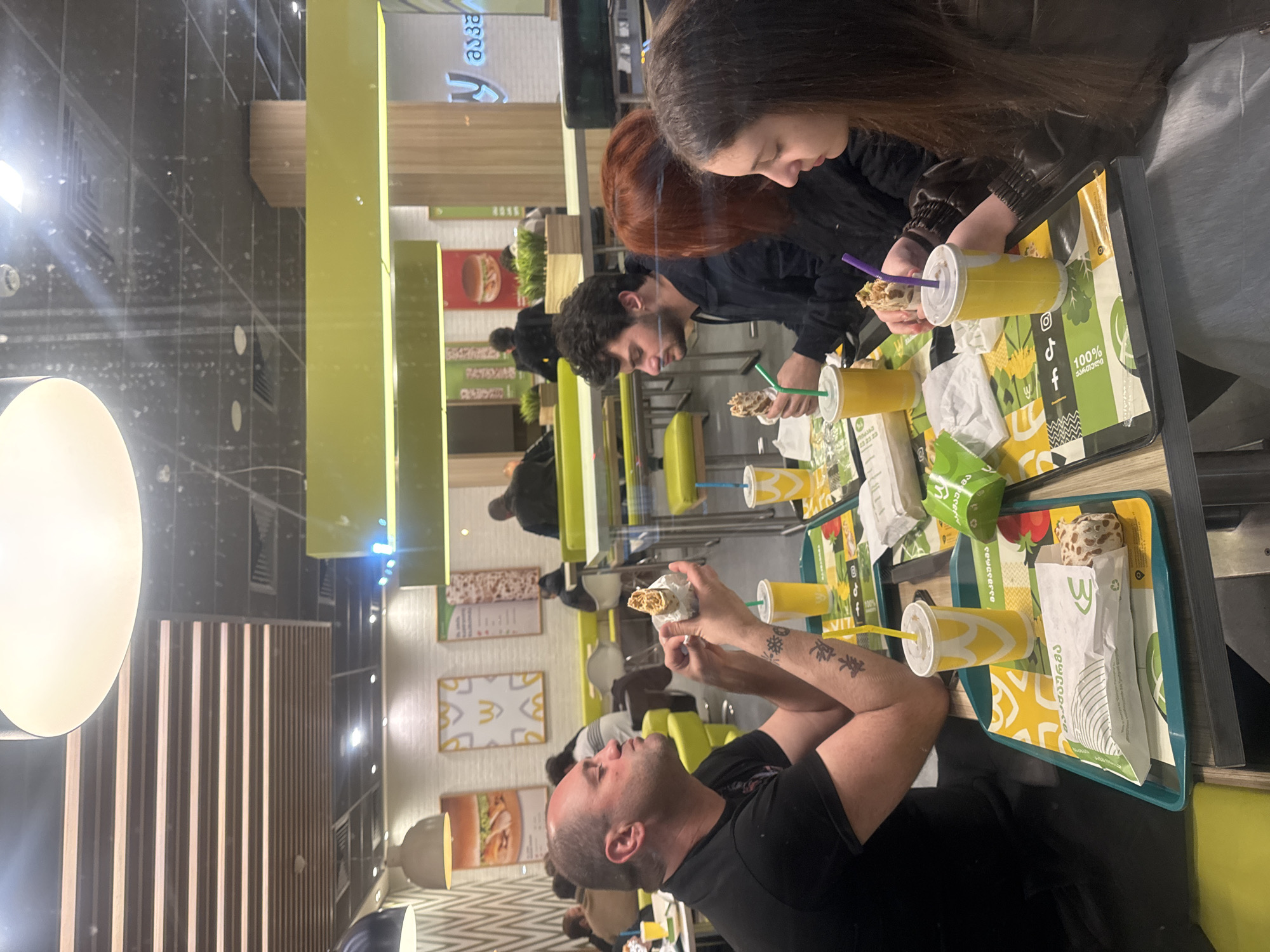 The image shows a group of people dining in a McDonald's restaurant. The location is likely in Tbilisi, Georgia, based on the "100% ქართული" text on the packaging, which translates to "100% Georgian".
There are at least four people visible: a woman with brown hair, a woman with red hair, a man with curly dark hair, and a bald man with tattoos on his arms. They are sitting at a table, holding and eating food items wrapped in paper.
Each person has a yellow cup with a straw, presumably containing a beverage. The food appears to be wrapped sandwiches or wraps. The food and drinks are placed on trays.
The background shows the interior of the McDonald's restaurant with yellow and white color schemes. There are posters and signs on the wall, including images of burgers. Other customers are seated at tables in the background. The floor is dark, and a lamp is visible on the left.