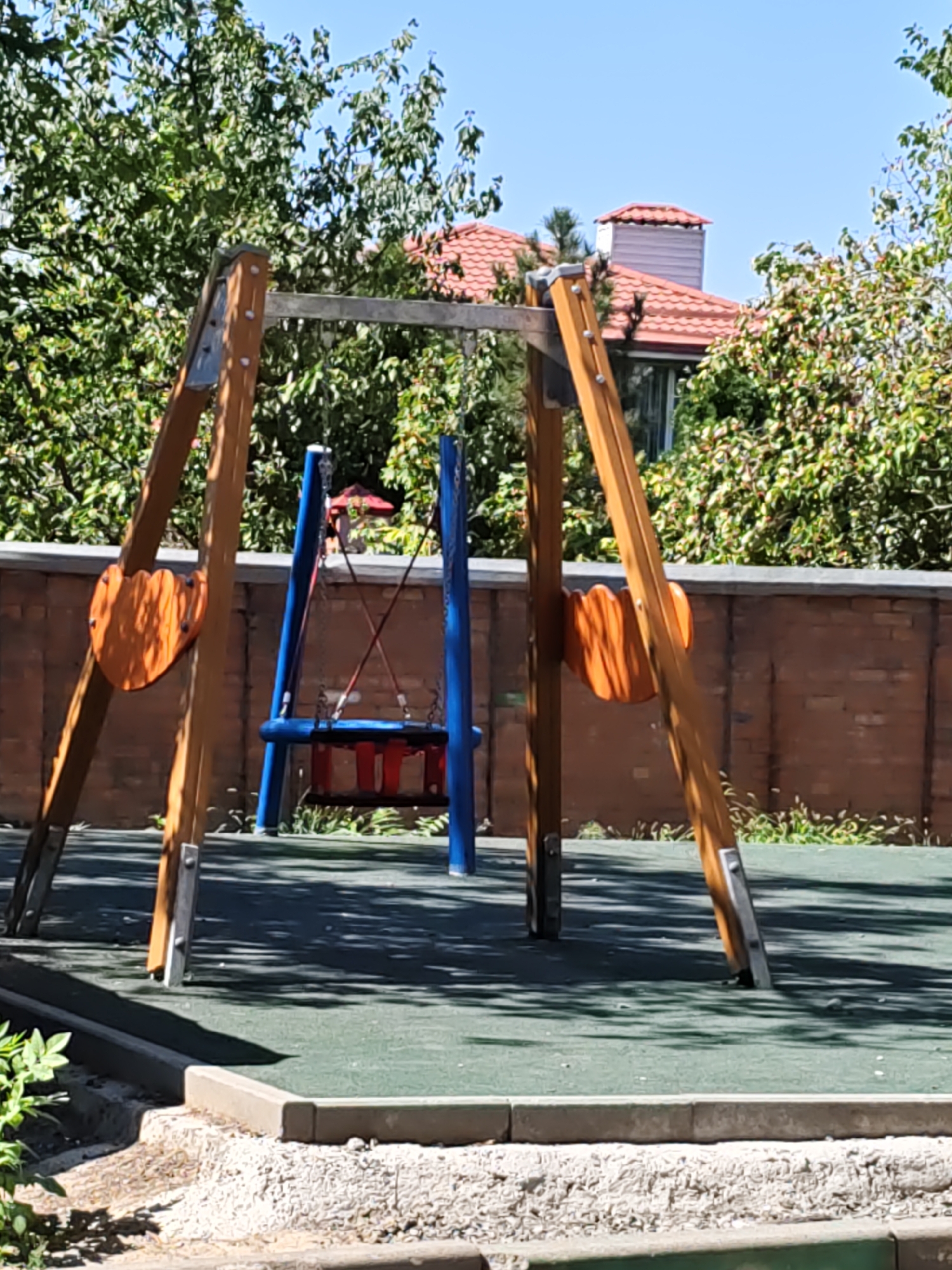 The image shows an outdoor playground with a swing set made of wooden beams and blue metal supports. A red and blue swing seat is suspended from the frame. In the background, a brick wall is visible, with trees and a residential building with a red-tiled roof behind it. The playground surface appears to be green rubber matting. The scene is bathed in sunlight, casting shadows from the swing set onto the ground. The location is identified as a playground in Rostov-on-Don, Russia. No people are present in the image.