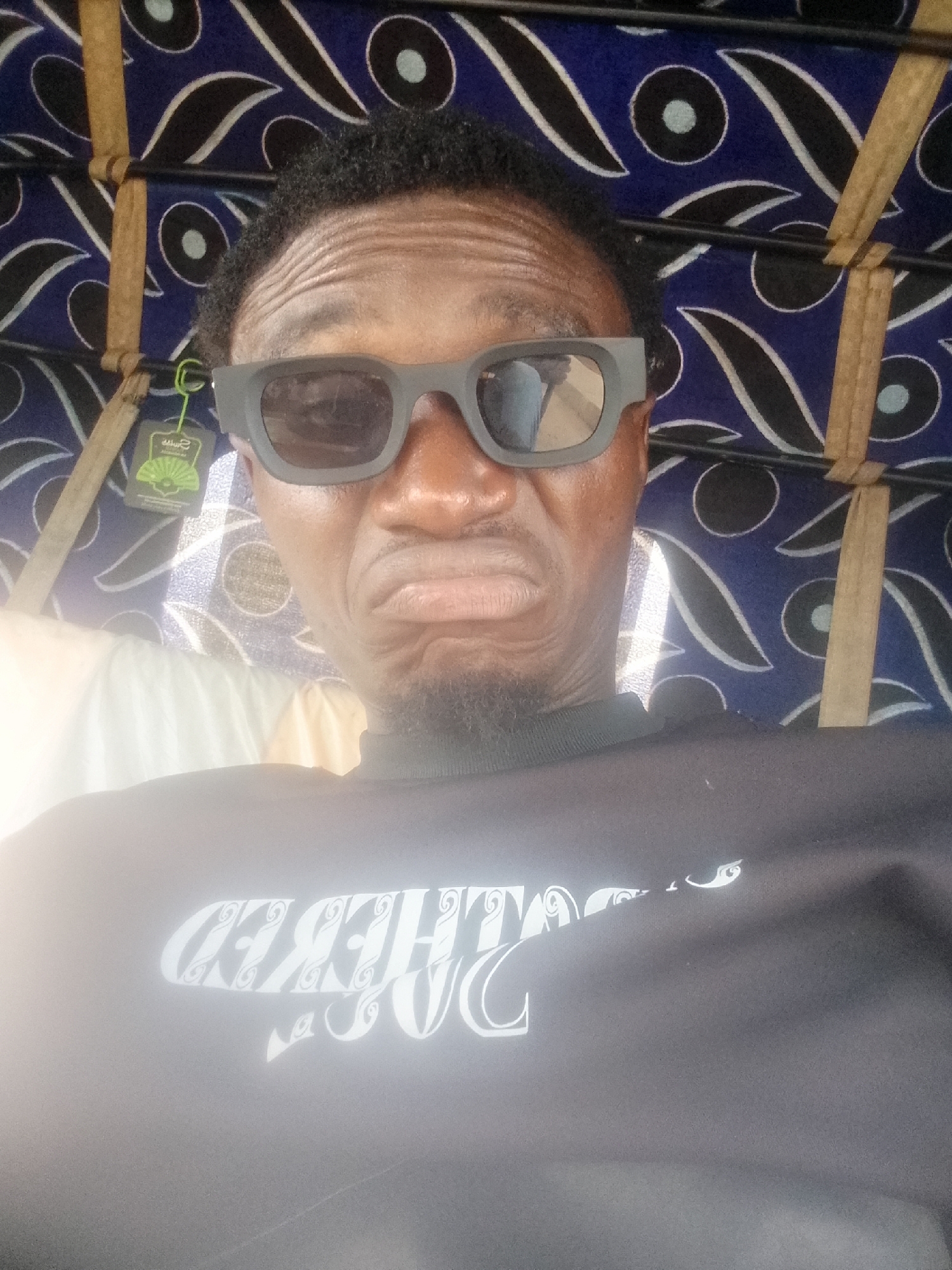 A selfie taken by a dark-skinned man wearing dark, wide-framed sunglasses and a black turtleneck sweatshirt with light-colored text, possibly in Bukuru, Nigeria. The man is frowning and looking into the camera. The background features a blue patterned fabric, likely part of a vehicle or shelter. Tan wooden slats are also visible in the background. A green price tag hangs from the sunglasses.