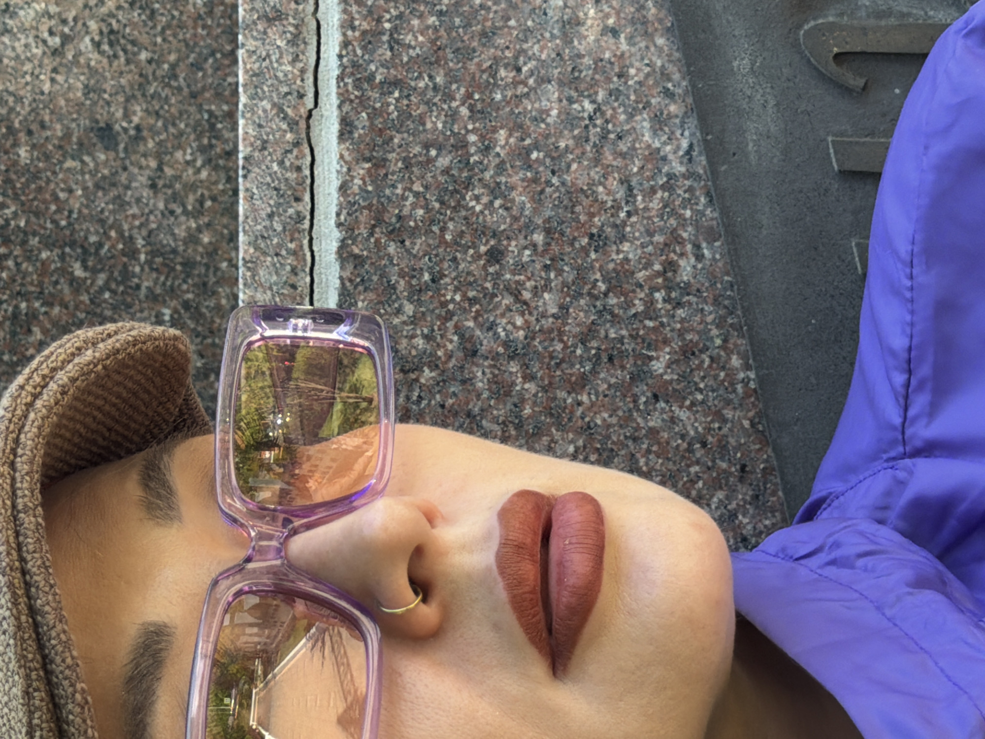 A person is lying down with their head resting on a granite surface, possibly a monument or sidewalk. They are wearing a brown knit cap, sunglasses with pink frames, a gold nose ring, and a purple jacket or coat. The sunglasses reflect a park-like scene. The surface beside the person's head is made of granite with a crack or seam visible. There is a possible partial letter etched in a dark stone beside the person, suggesting they are near a memorial or grave marker. There is insufficient information to identify the precise location as being in Krasnoyarsk.