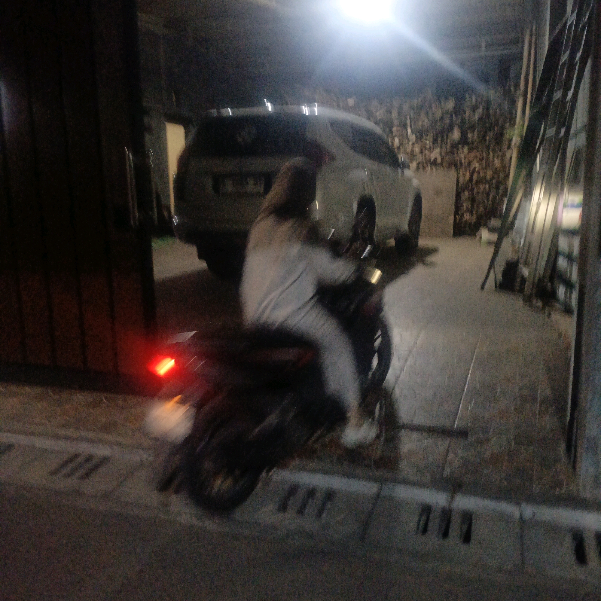 The image depicts a scene at night, likely in an outdoor, possibly covered or semi-enclosed, area. A person is seated on a dark-colored motorcycle, wearing light-colored clothing, possibly a jacket and pants. The motorcycle's taillight is illuminated with a bright red glow. In the background, a white SUV is partially visible, facing towards the left of the frame. The environment appears to be a paved area with some indistinct structures and objects to the right, including what seems to be scaffolding or tall, narrow objects. The lighting is dim, with a bright light source emanating from the upper left, creating glare. The image quality is poor due to motion blur and low light conditions. The scene does not contain any immediately identifiable landmarks or specific features that would definitively place it in Salatiga, Indonesia, or any other specific city without further context.