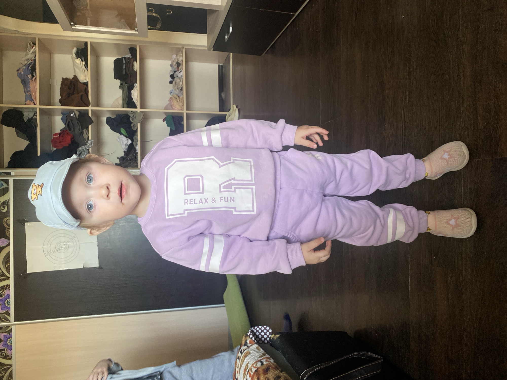 A child is lying on their side on a dark wood floor in a room with a wooden storage cabinet in the background. The child is wearing a light purple tracksuit with white stripes on the sleeves and legs. The words "RELAX & FUN" are printed on the chest. The child is also wearing a light blue baseball cap with a cartoon image on the front and light pink shoes. A target is affixed to a dark-colored surface near the child's head. Part of a couch is visible at the bottom of the frame.
The image was taken in Chaikovsky, Russia.