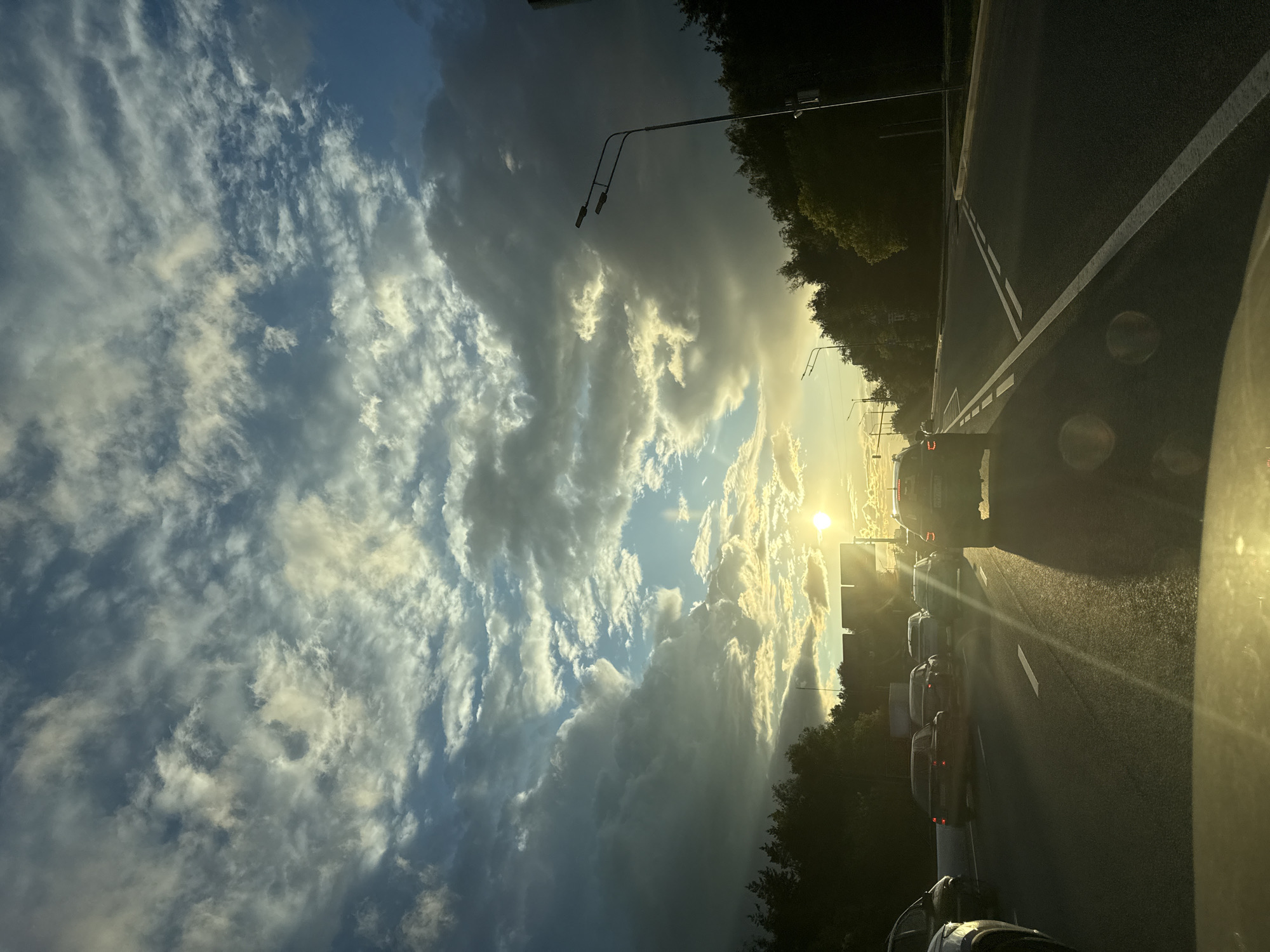 The image depicts a road in Moscow, Russia at sunset, with cars lined up in traffic. The sky is visible with clouds illuminated by the setting sun. On both sides of the road are trees and street lights.
