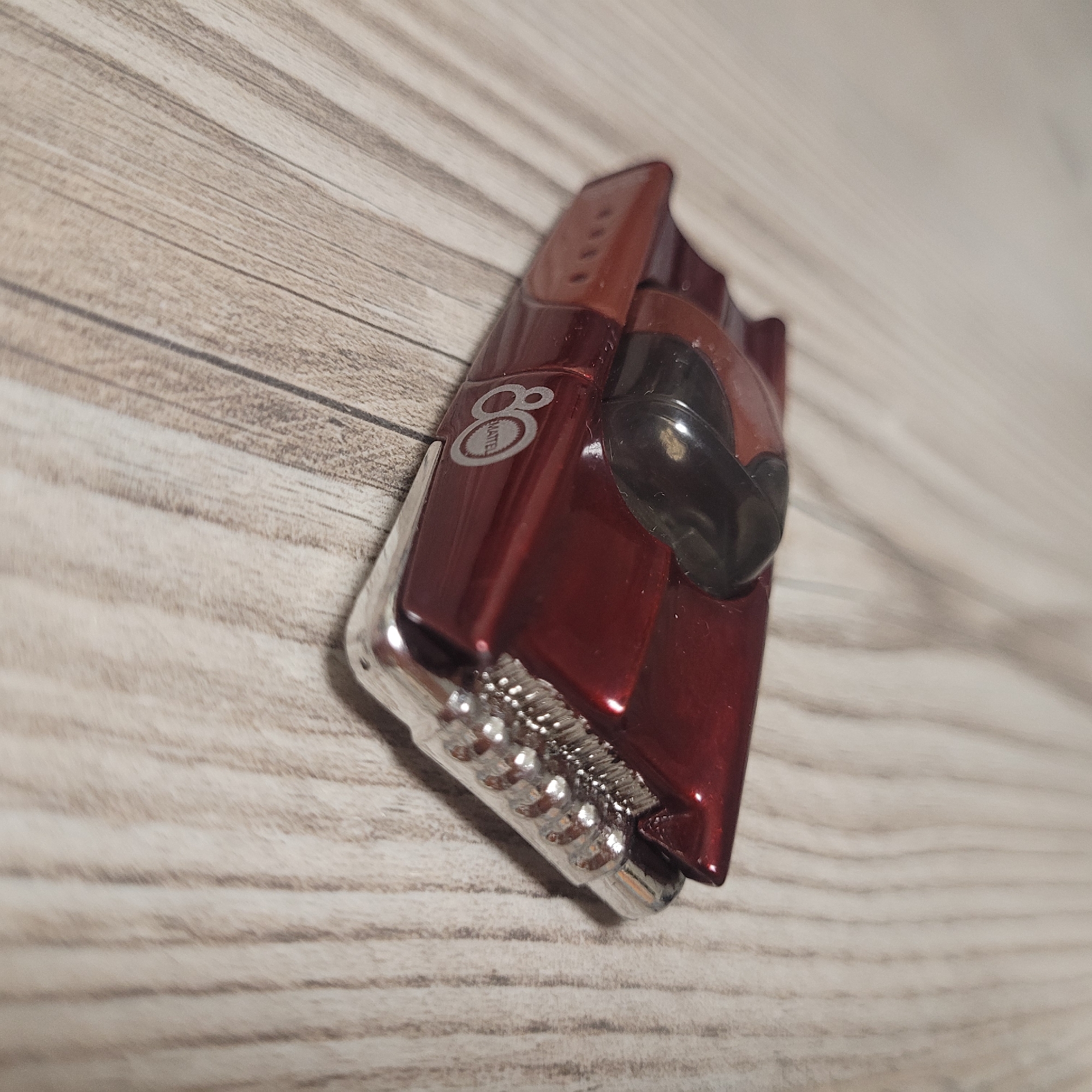 The image shows a close-up, angled view of a metallic red electric shaver with a black plastic head. The shaver is lying on a wooden surface with a light, natural wood grain pattern. The shaver has a sleek, modern design, featuring a glossy finish. On the side of the shaver, there is a circular logo with the number "8" and the word "AMATELL" inside. The shaver has a row of metallic teeth at the bottom, suggesting it's a trimmer or grooming device. The lighting is overhead, casting a slight shadow on the wooden surface. The overall impression is of a product shot, focusing on the details of the grooming tool. There are no people or other objects visible in the frame. The setting appears to be indoors, on a tabletop or desk.