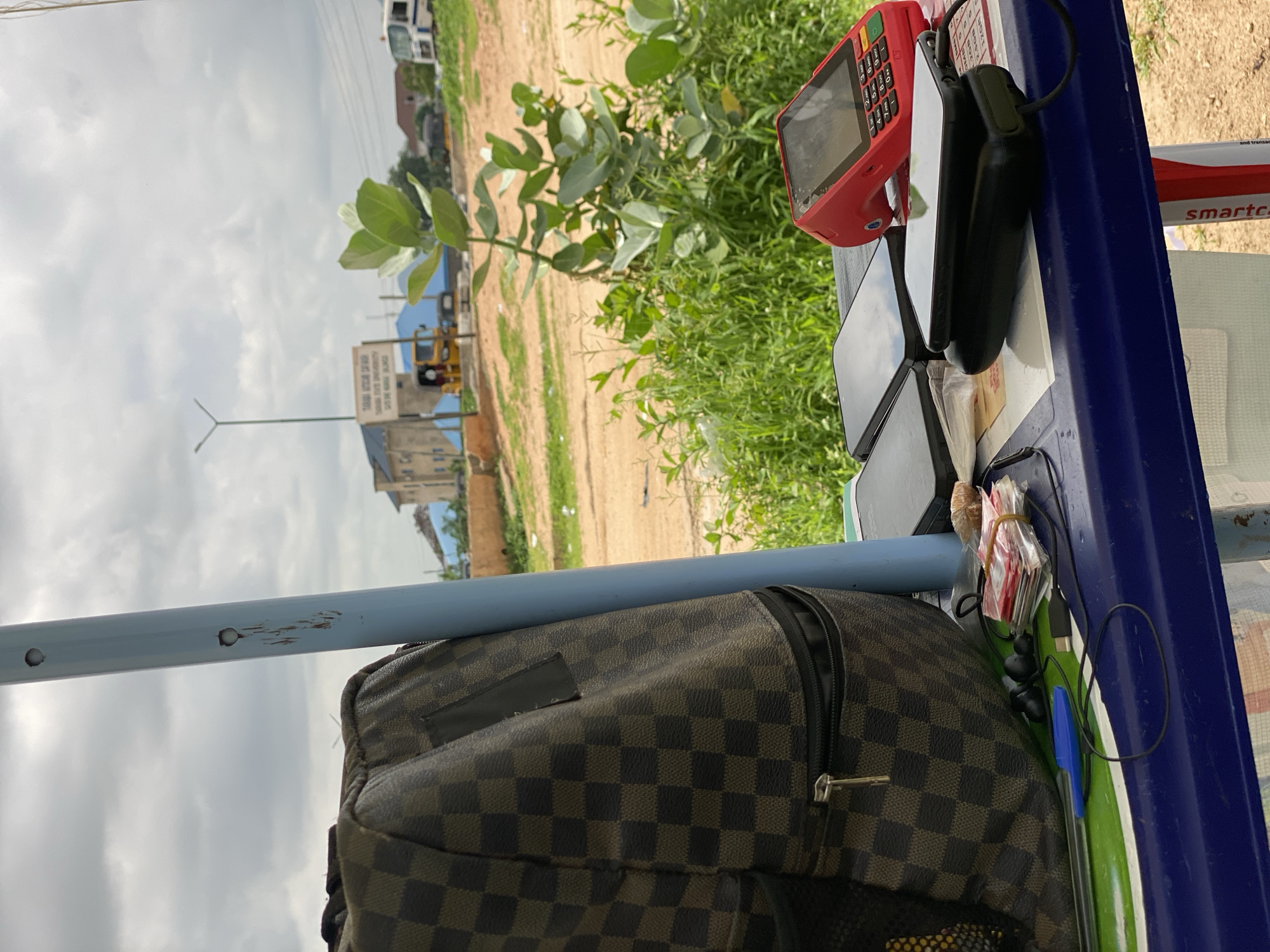 The image depicts an outdoor scene in Jalingo, Nigeria, likely near a roadside stall. In the foreground, a dark checkered backpack is positioned to the left, resting on a blue surface that appears to be a table or counter. Several electronic devices are laid out on this surface, including a red point-of-sale (POS) terminal, two black smartphones, and a black power bank. A blue stylus pen, a pair of black earbuds, and a bundle of cash secured with a rubber band are also visible.
In the mid-ground, lush green vegetation and a young tree with large leaves dominate the scene. Beyond the foliage, a dirt road is visible, with a yellow commercial tricycle (keke napep) and other vehicles in the distance. A signpost with text, partially obscured, is also present. The background shows buildings and a cloudy sky. The overall environment suggests a commercial area or a rest stop in Jalingo. No people are clearly visible in the image, but the arrangement of items suggests a commercial transaction or waiting period.