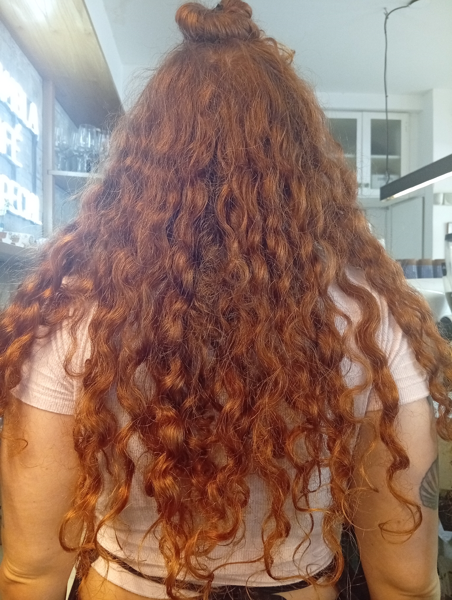 A woman is seen from the back, her long, curly, auburn hair pulled into a high bun. She is wearing a light pink, short-sleeved shirt, and the top of her black bottoms is visible. She has a tattoo on her upper left arm. The background includes a wooden shelf with glasses, white cabinets, and a modern, linear light fixture. The location is inside a building, possibly in Buenos Aires, Argentina, but there is no direct confirmation.