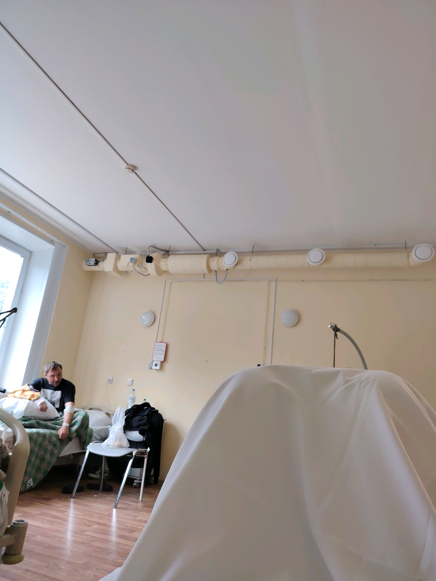The image depicts a hospital room in Marino, Russia. A man with a bandage on his left arm is seated on a hospital bed. The room features a window, a chair, medical equipment, and various supplies. A large white sheet is draped over an object in the foreground, obscuring its identity. The overall atmosphere suggests a medical facility.