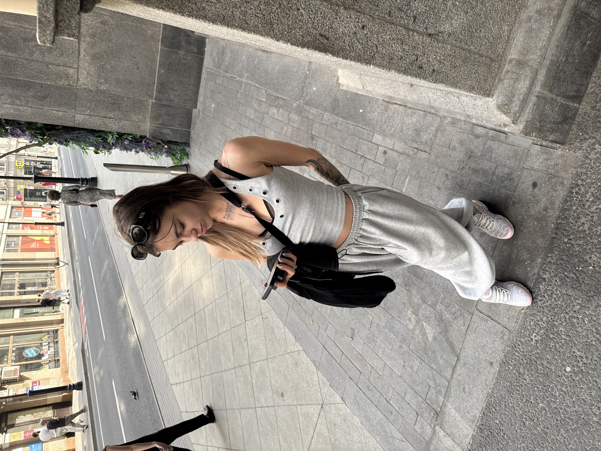 A young woman with blonde hair, wearing sunglasses on her head, a grey tank top, grey sweatpants, and athletic shoes, stands on a grey stone sidewalk, looking at her phone. She is holding a black jacket over her left arm. She has tattoos on her arms and chest. Behind her, a street with cars and pedestrians is visible. The scene appears to be in Moscow, Russia, given the architectural style and the presence of Cyrillic signage on buildings in the background. The lighting suggests it is daytime, likely afternoon.