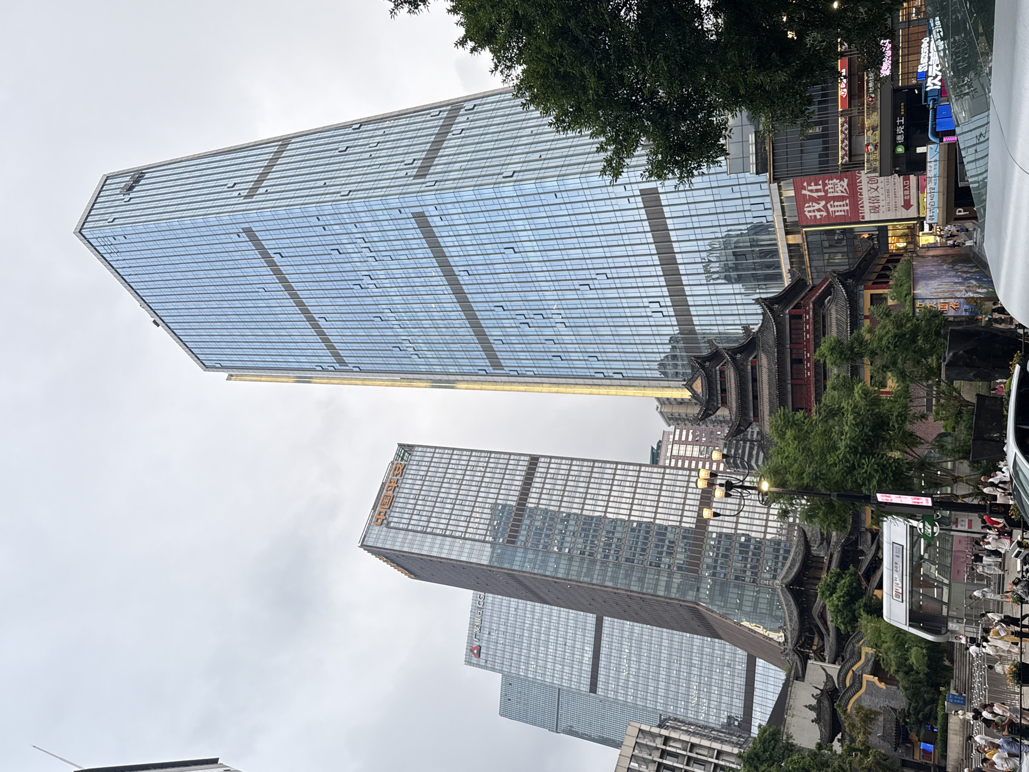 The image depicts a street scene in Yuzhong District, China, characterized by a striking juxtaposition of modern skyscrapers and traditional Chinese architecture. A towering, blue-glass skyscraper dominates the right side of the frame, its facade reflecting the overcast sky. To its left, two other skyscrapers of similar modern design are visible, one bearing the orange text "中国平安" (Ping An Insurance) and another with a red triangle logo and the text "广发银行" (Guangfa Bank).
In the foreground and mid-ground, a cluster of traditional Chinese buildings with dark, curved roofs and ornate eaves provides a stark contrast to the contemporary structures. These buildings appear to be part of a temple or a historic complex. A busy street is present, with numerous people walking and gathered. A white metro station entrance with Chinese signage is visible in the lower center of the image, suggesting an active urban environment. Several trees with lush green foliage are interspersed throughout the scene, softening the urban landscape. A street lamp with multiple globes stands near the traditional buildings. A red banner with Chinese characters, including "我在重庆" (I am in Chongqing), is prominently displayed on one of the buildings. The overall atmosphere is that of a vibrant city where historical and modern elements coexist.