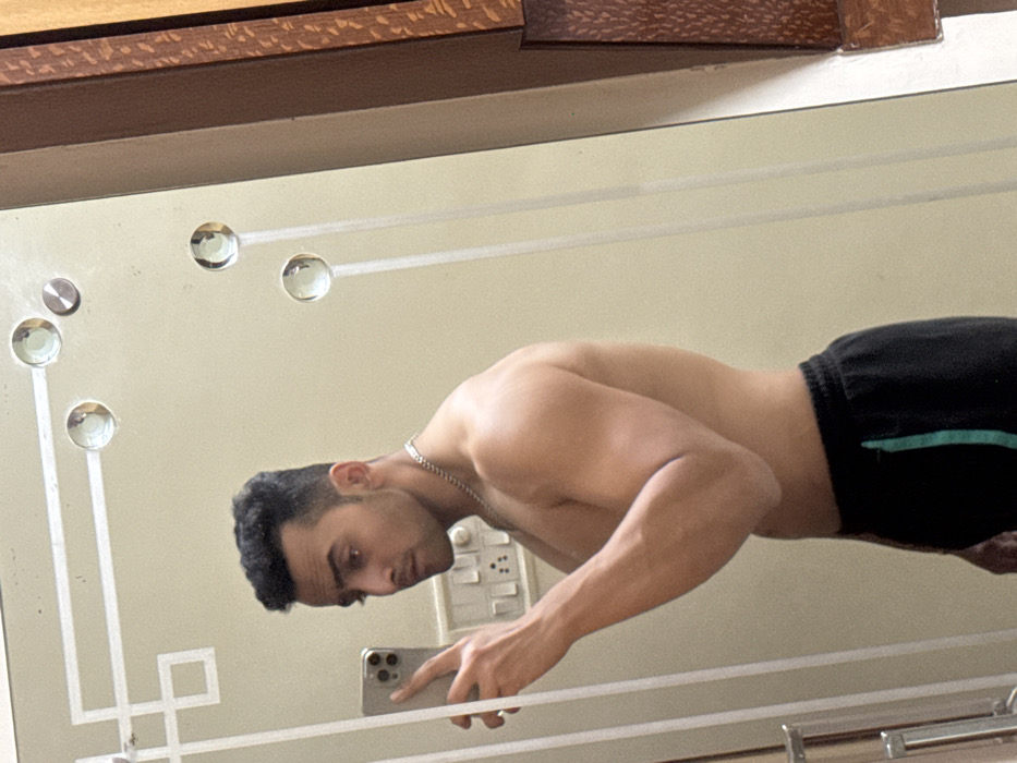 A shirtless man is seen in a mirror selfie. He is wearing black athletic shorts with a green stripe on the side. His head is turned to the left, and he is looking at his phone. He has short, dark hair. The mirror has a decorative design of white lines and circles. The man is also wearing a silver necklace. In the background, there is a light switch. The reflection is angled.