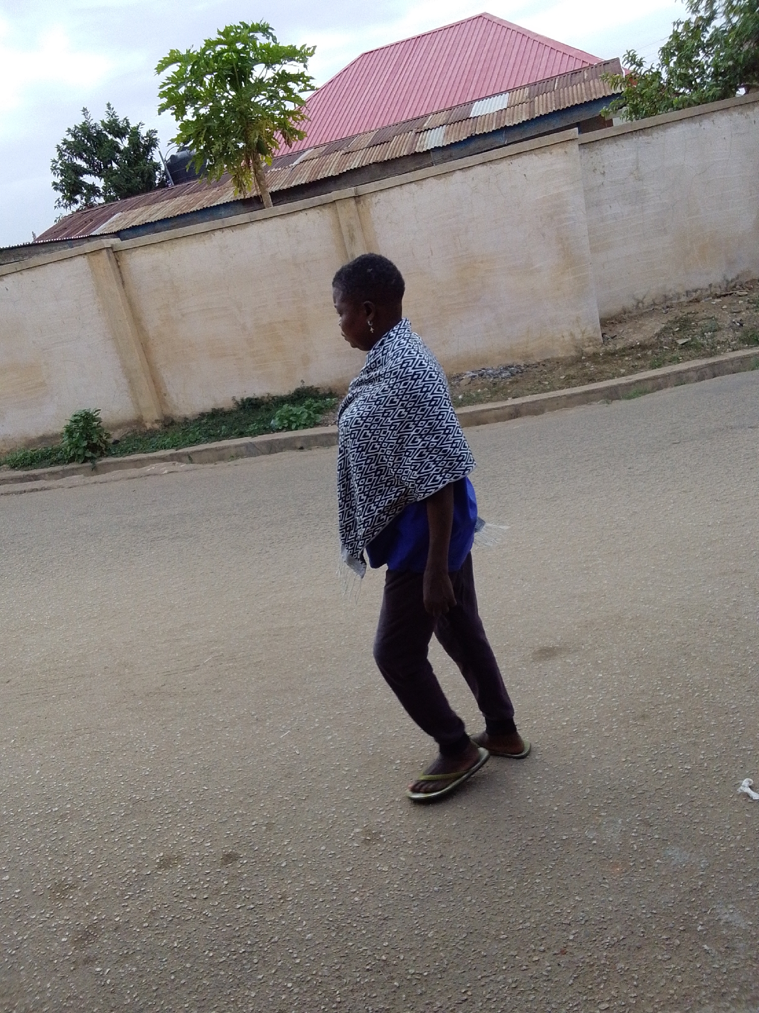 An individual, likely an adult female, is captured walking on a paved road, viewed from her right side as she moves towards the right of the frame. She has short, dark, curly hair and wears a blue shirt, dark trousers, and olive green flip-flops. A distinctive black and white geometric patterned shawl or wrap covers her shoulders. A small earring is visible on her right ear.
The background features a long, light-colored concrete wall, likely a perimeter fence. Behind this wall, various buildings are visible, characterized by corrugated metal roofs; some appear rusty, while others have a distinct reddish color. A black water tank is partially visible on one of the roofs. Above the structures, lush green vegetation includes a prominent papaya tree, which appears to be bearing fruit, and other dense green trees, suggesting a tropical or subtropical environment.
The sky is uniformly light grey and overcast, indicating either early morning, late afternoon, or a cloudy day, with diffused lighting across the scene. The road in the foreground and midground is a light grey, coarse asphalt surface. A narrow strip of ground with minimal greenery and a small curb separates the road from the concrete wall. A small piece of white litter is visible on the road surface in the bottom right corner. No visible text is present in the image. The scene depicts a mundane moment in a street environment, consistent with the specified location of Jos, Nigeria.