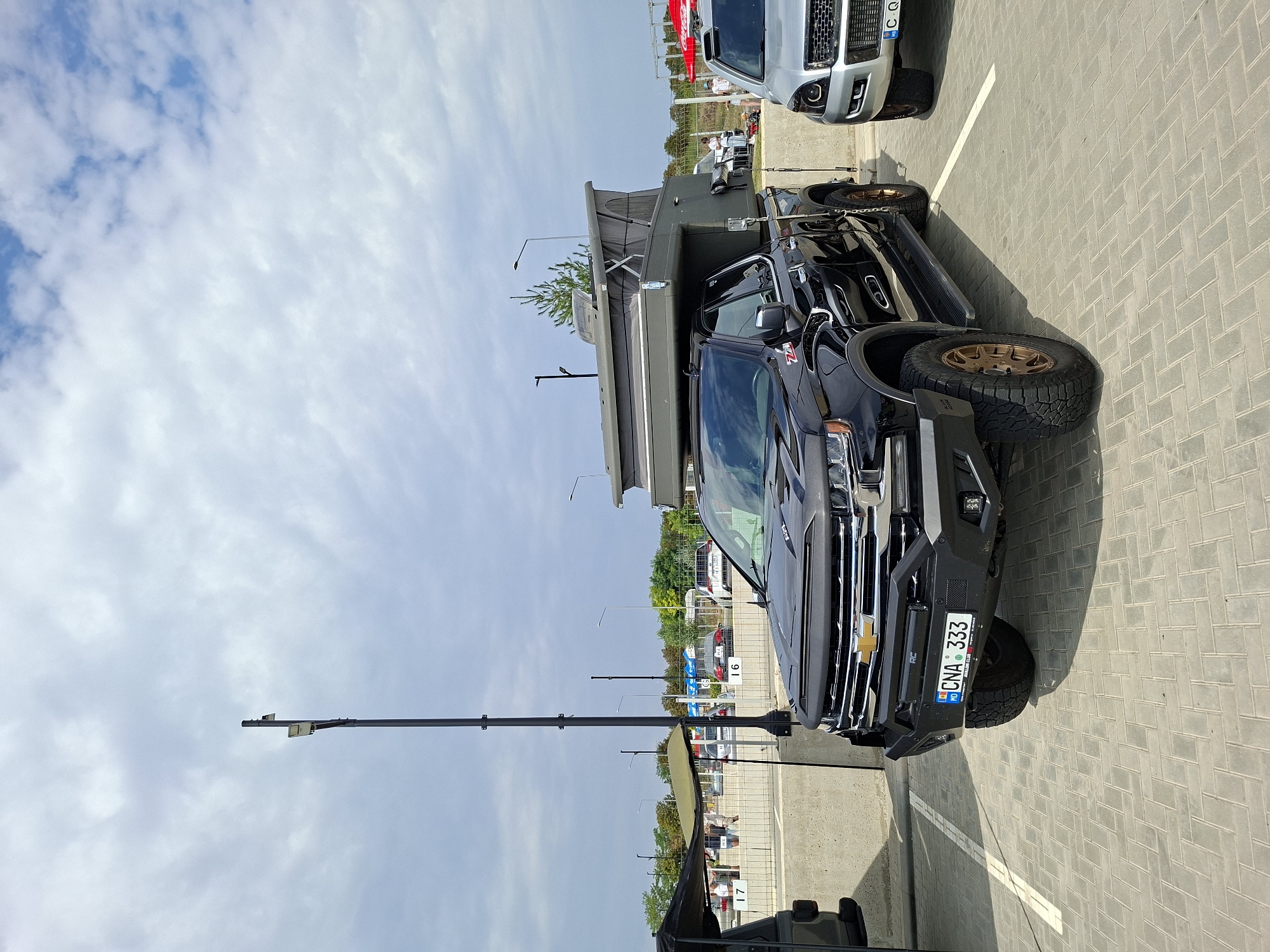 A black Chevrolet Silverado pickup truck is parked in an outdoor lot. The truck is equipped with a pop-up camper on its bed. It has a modified front bumper with lights and a license plate reading "MD CNA 333". The truck is parked next to a silver SUV. In the background, there is a concrete barrier with parking spot numbers and trees. A lamppost with two lights is visible to the left of the truck. The sky is cloudy. The scene appears to be a parking area, potentially at an event or a campground, located in Stăuceni, Moldova.