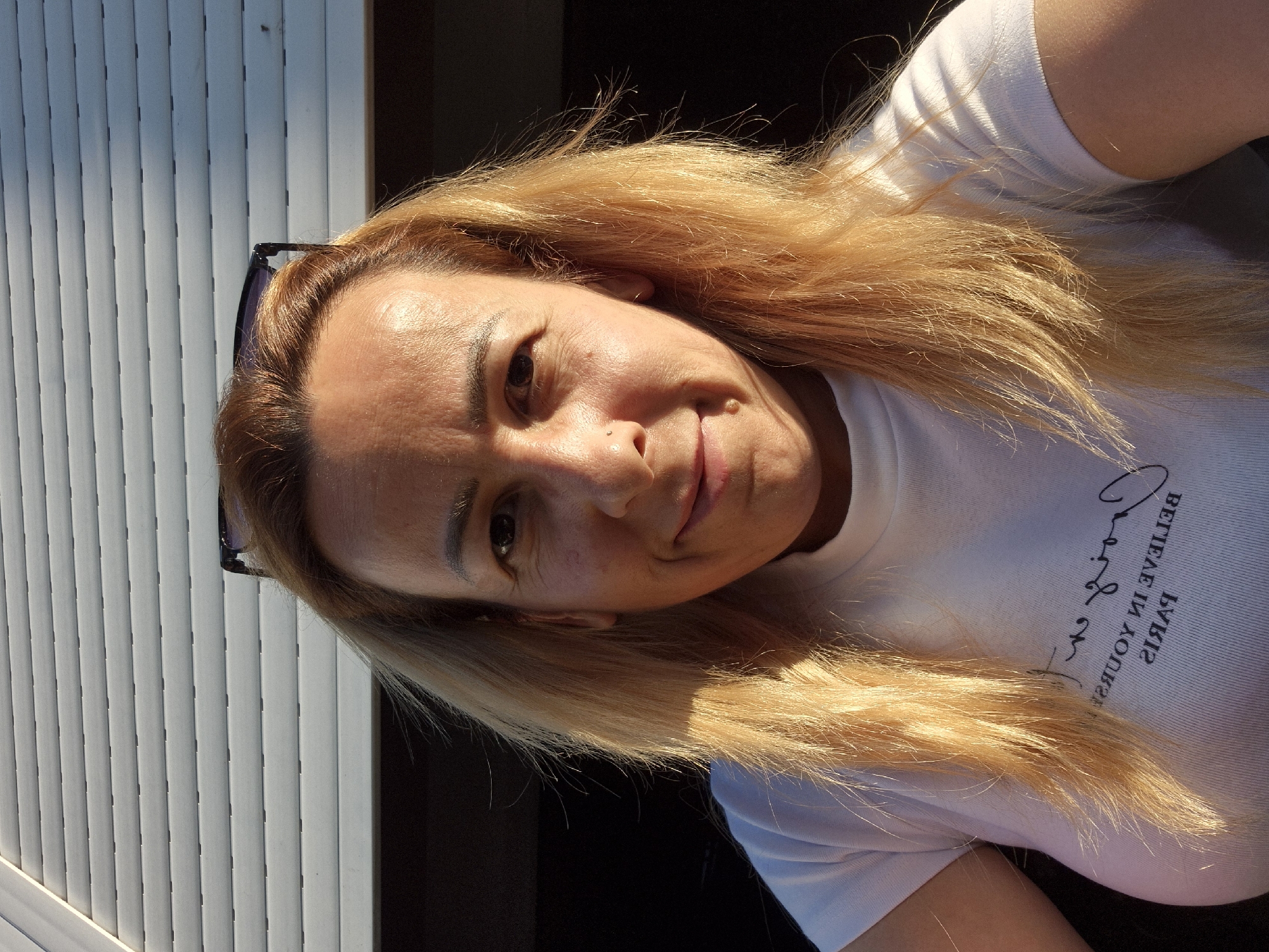 This is a selfie of a woman with blonde hair. She is wearing a white t-shirt with text on it. The text on her shirt reads "Believe in yourself Paris" in cursive writing. She is wearing sunglasses on her head. The background consists of white blinds and a dark area. The lighting suggests it is daytime, possibly late afternoon due to the warm tones of the light on her hair. The woman's expression is a slight smile.
