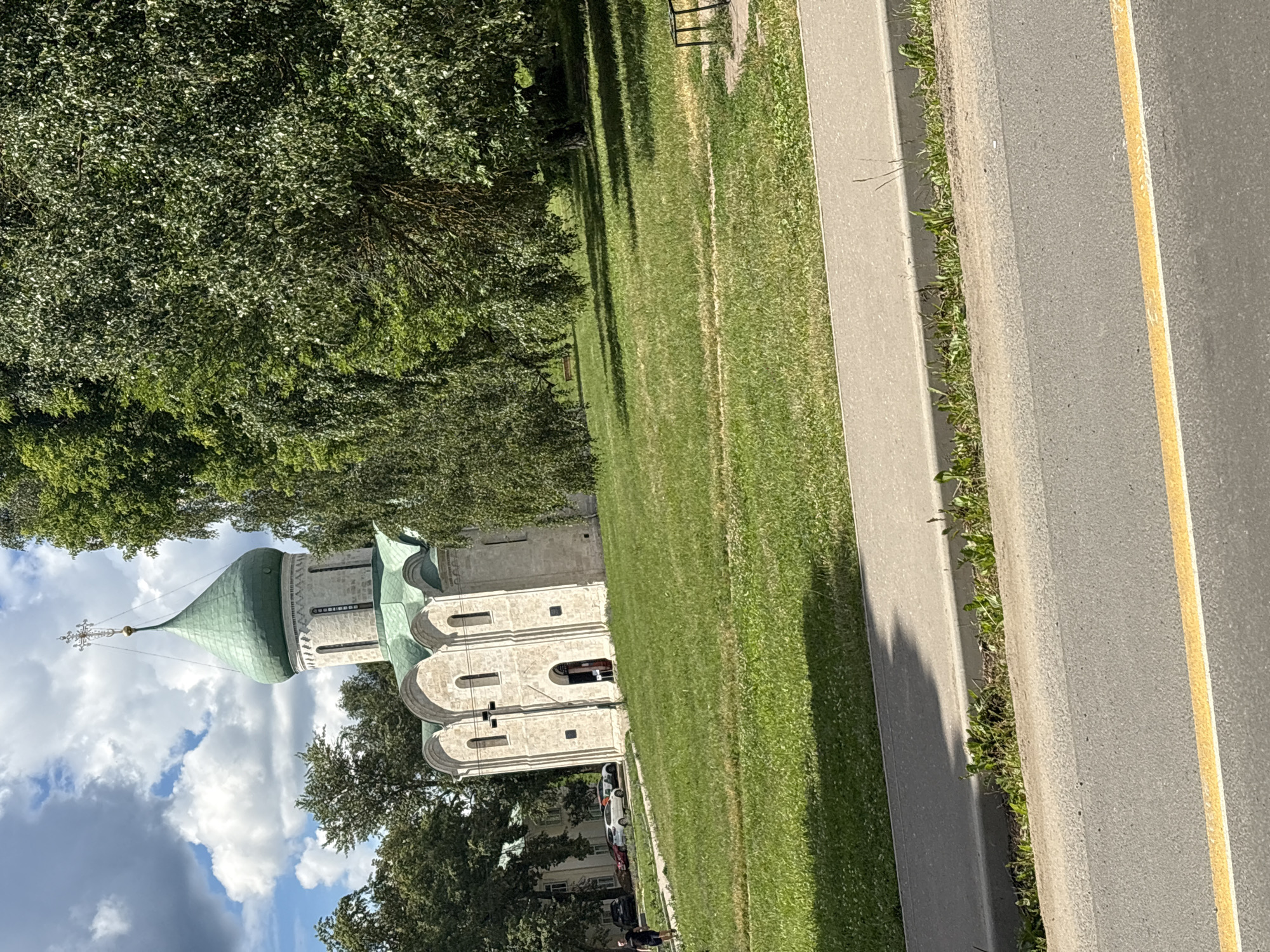The image shows the Church of St. Theodore the Black in Pereslavl-Zalessky, Russia, on a sunny day. The white stone church has a green, onion-shaped dome topped with a cross. It is surrounded by a well-maintained green lawn and mature trees. A paved sidewalk runs along the foreground, with a road beyond it. In the background, several cars are parked near another building. A person in dark clothing is walking on the lawn. The sky is blue with scattered white clouds.