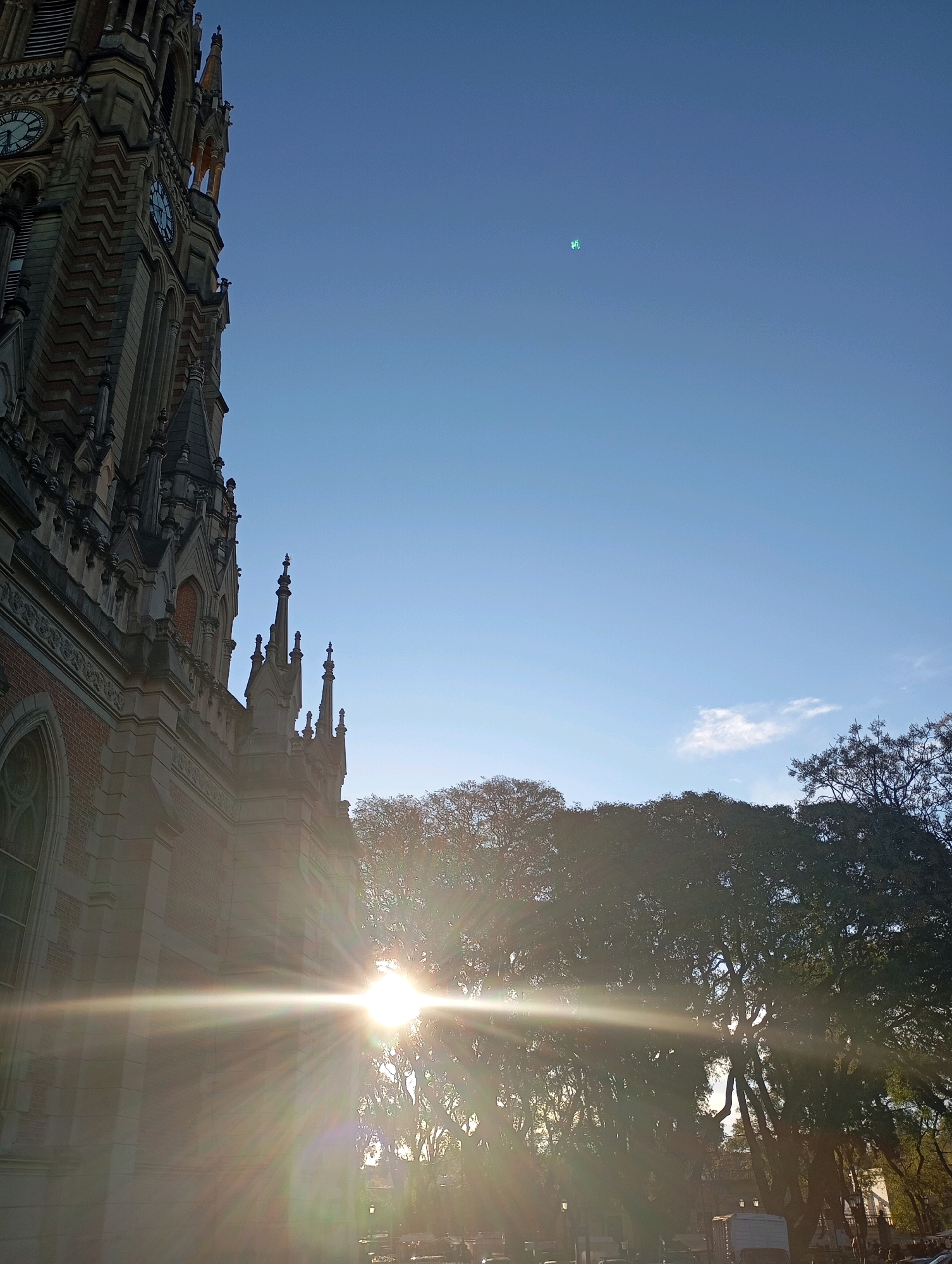 The image depicts a section of the Cathedral of San Isidro, a prominent landmark in the city of San Isidro, Argentina, on a bright, clear day. The cathedral's Gothic-style architecture, characterized by its pointed arches, spires, and intricate stonework, is visible on the left side of the frame, bathed in the warm glow of the sun.
The sun, positioned in the mid-right of the image, casts a strong lens flare across the scene, creating brilliant streaks of light that emanate from its source. The light filters through the dense foliage of mature trees, silhouetting their branches and creating a dappled effect. The trees line a street or plaza, with glimpses of lampposts and what appear to be vehicles and a small white structure in the background, suggesting an urban setting.
The sky above is a clear, pale blue, with a faint hint of a small, wispy cloud in the upper right quadrant. There are no discernible people in the image, nor any direct interactions taking place. The focus is on the interplay of light and shadow, architecture, and nature within the urban landscape of San Isidro. The overall impression is one of a serene and sun-drenched afternoon in this Argentine city.