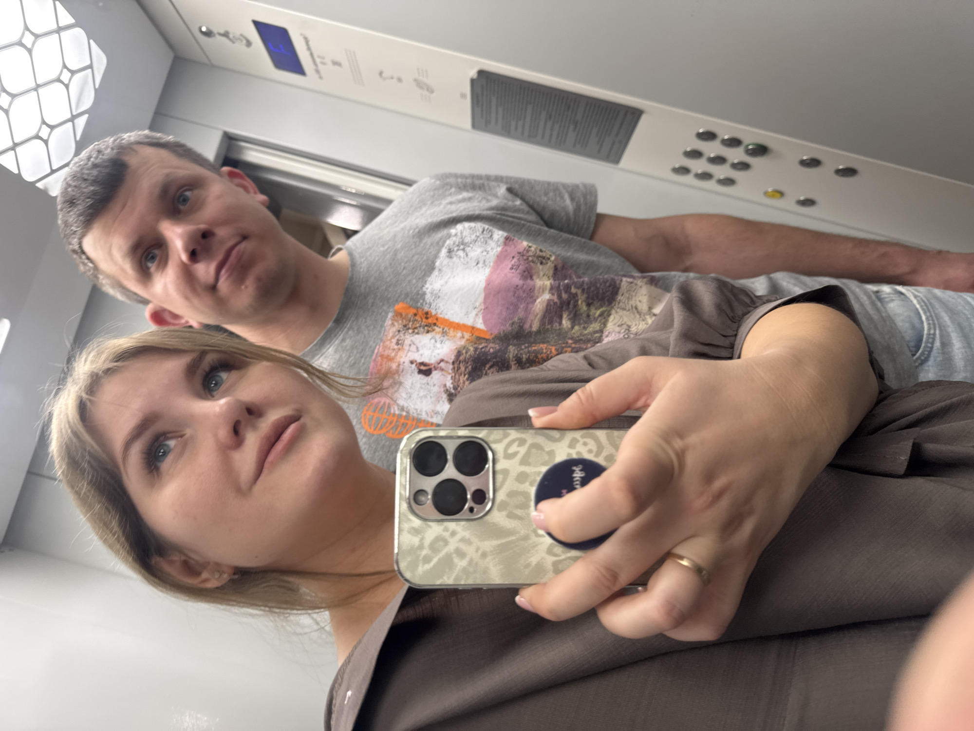A selfie taken in an elevator. A woman is holding a phone with a case featuring a leopard print design and a black pop socket. She has light blonde hair and is wearing a brown top. A gold ring is visible on her finger. Behind her stands a man with short, graying hair and a gray T-shirt with a graphic print. The elevator's interior has a control panel with buttons and a digital display. The location is indicated as the city of Chaikovsky, Russia.
