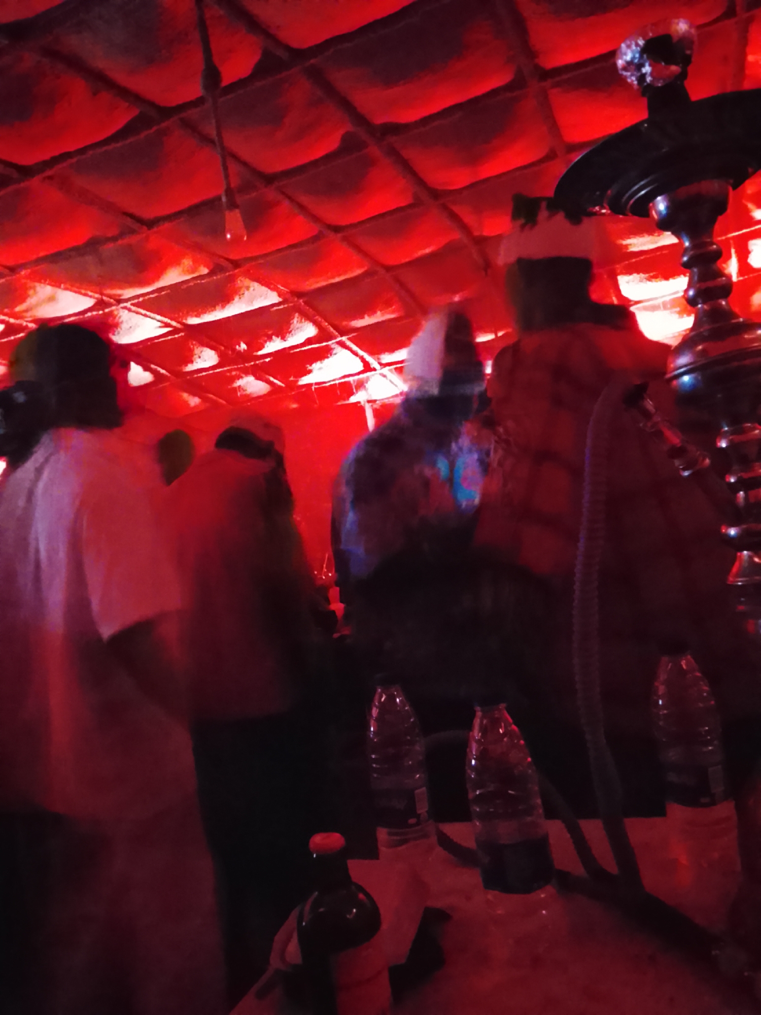 The image depicts a dimly lit interior scene, possibly a lounge or recreational space, illuminated by vibrant red lighting that casts a strong glow on the surroundings. In the foreground, a cluster of plastic water bottles is visible, some appearing to be partially filled. One bottle is placed on a surface, with a dark-colored bottle with a red cap situated in front of it. To the right, the metallic stem and base of a hookah pipe are discernible, suggesting the presence of shisha smoking facilities.
Further back, several individuals are present. On the left, a person wearing a white shirt is partially visible from the side, their face obscured by shadow and the red lighting. To their right, another individual in a patterned shirt and a white head covering stands. Further to the right, a person in a red and black plaid shirt and a white head covering is observed from the side. The background features a textured ceiling with a grid-like pattern, also bathed in the red light. The overall ambiance suggests a social gathering in a place that embraces a modern, dimly lit aesthetic, potentially found in entertainment venues within Abuja, Nigeria.