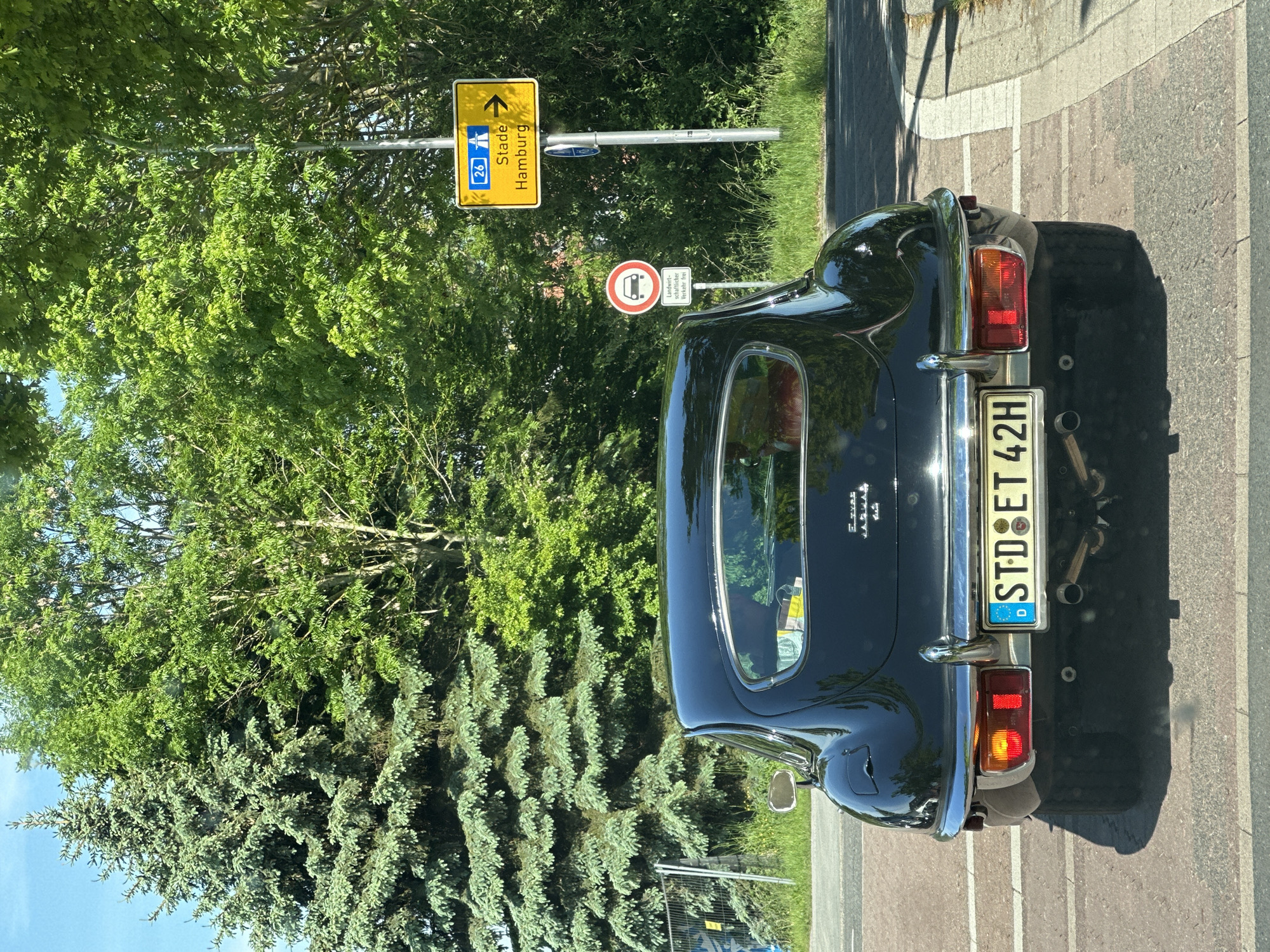 A dark-colored vintage Jaguar coupe is parked on the side of a road in Buxtehude, Germany, as indicated by the license plate "STD" (Stade district). The car is positioned near a road sign that points towards Stade and Hamburg (route 26). The sign for Landwirtschaftlicher Verkehr Frei (Agricultural traffic is allowed). The environment includes lush green trees, indicating a rural or suburban setting. The photo appears to have been taken on a sunny day.