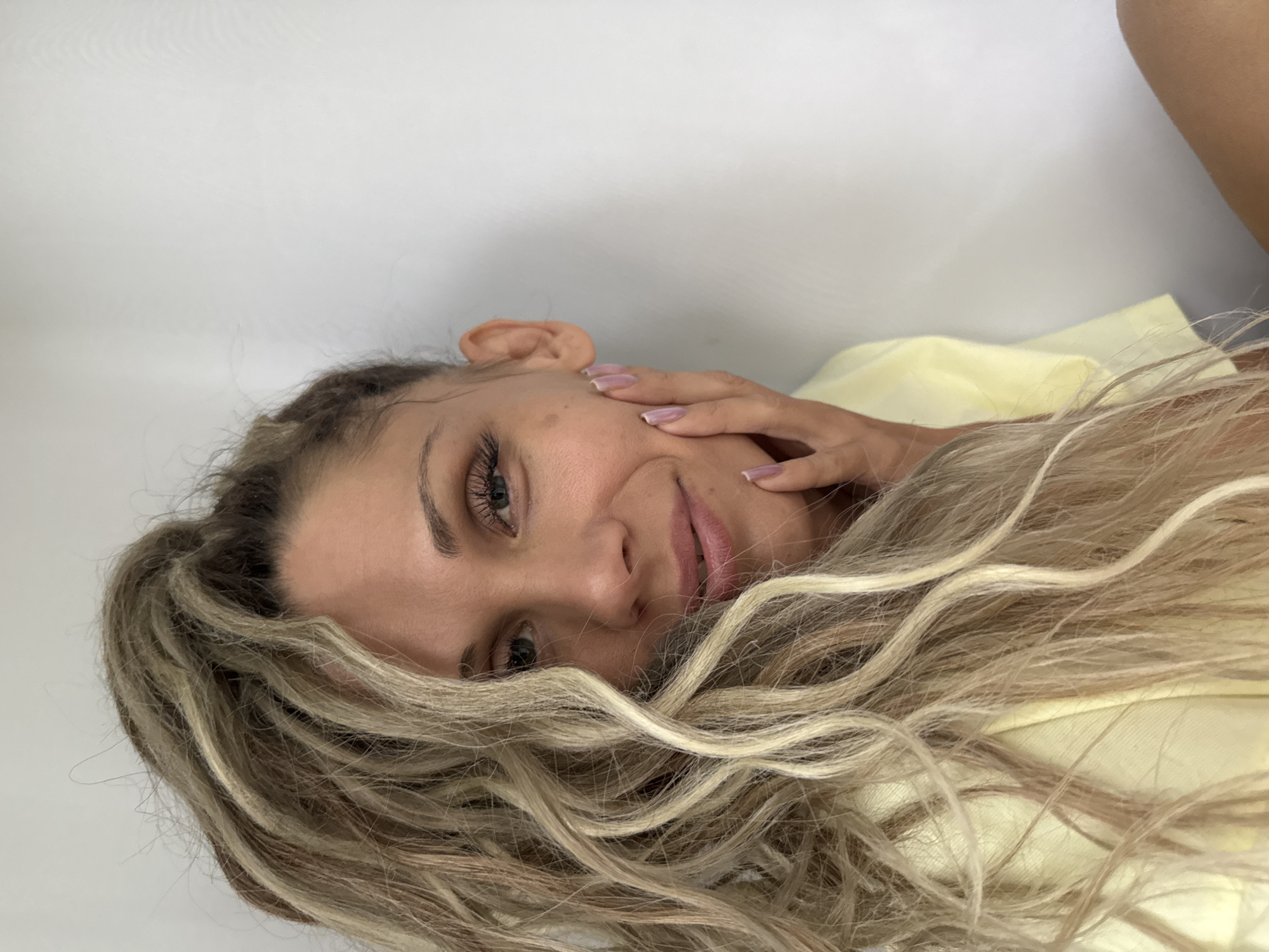 In this close-up, medium-shot photograph, a woman with long, wavy blonde hair is posed against a plain white background. Her hair is partially obscuring her face, with a few strands falling across her cheek and forehead. She is looking directly at the camera with a soft smile. Her right hand is resting on her left cheek, with her fingers gently touching her skin. She is wearing a pale yellow top. The lighting is soft and even, creating a natural and intimate feel. The location in the city of Zhukovo, Russia, is not discernible from the visual information provided in the image.
