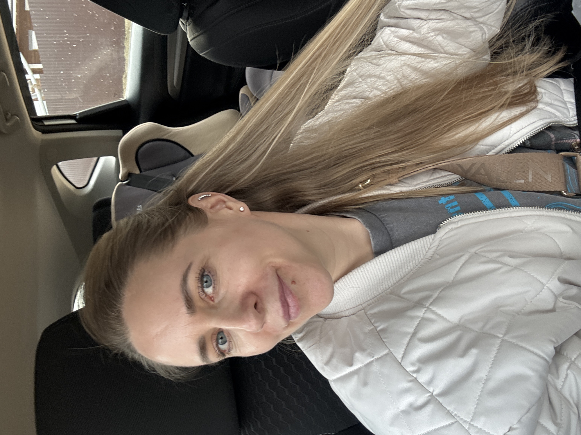 The image features a woman posing for a photo inside a car, situated in what is identified as Chaikovsky, Russia. She is positioned in the front passenger seat, looking directly at the camera with a gentle, pleasant smile.
The subject has long, blonde hair that falls over her shoulder, and striking blue eyes. A small mole is visible above her upper lip. She wears a light-colored, possibly beige or off-white, quilted jacket over a grey t-shirt with a partially visible blue graphic print. A beige strap, likely from a shoulder bag, crosses her body, displaying the text "VALN" which suggests a brand such as Valentino. Her left ear features multiple earrings: a small stud and another with a curved row of small stones.
The setting is clearly the interior of a vehicle, with dark fabric seats and the back of a child's car seat visible in the rear. Through the car window, an outdoor scene is discernible, showing what appears to be part of a building with vertical paneling, indicating a daytime setting. The woman's jacket suggests cooler weather.