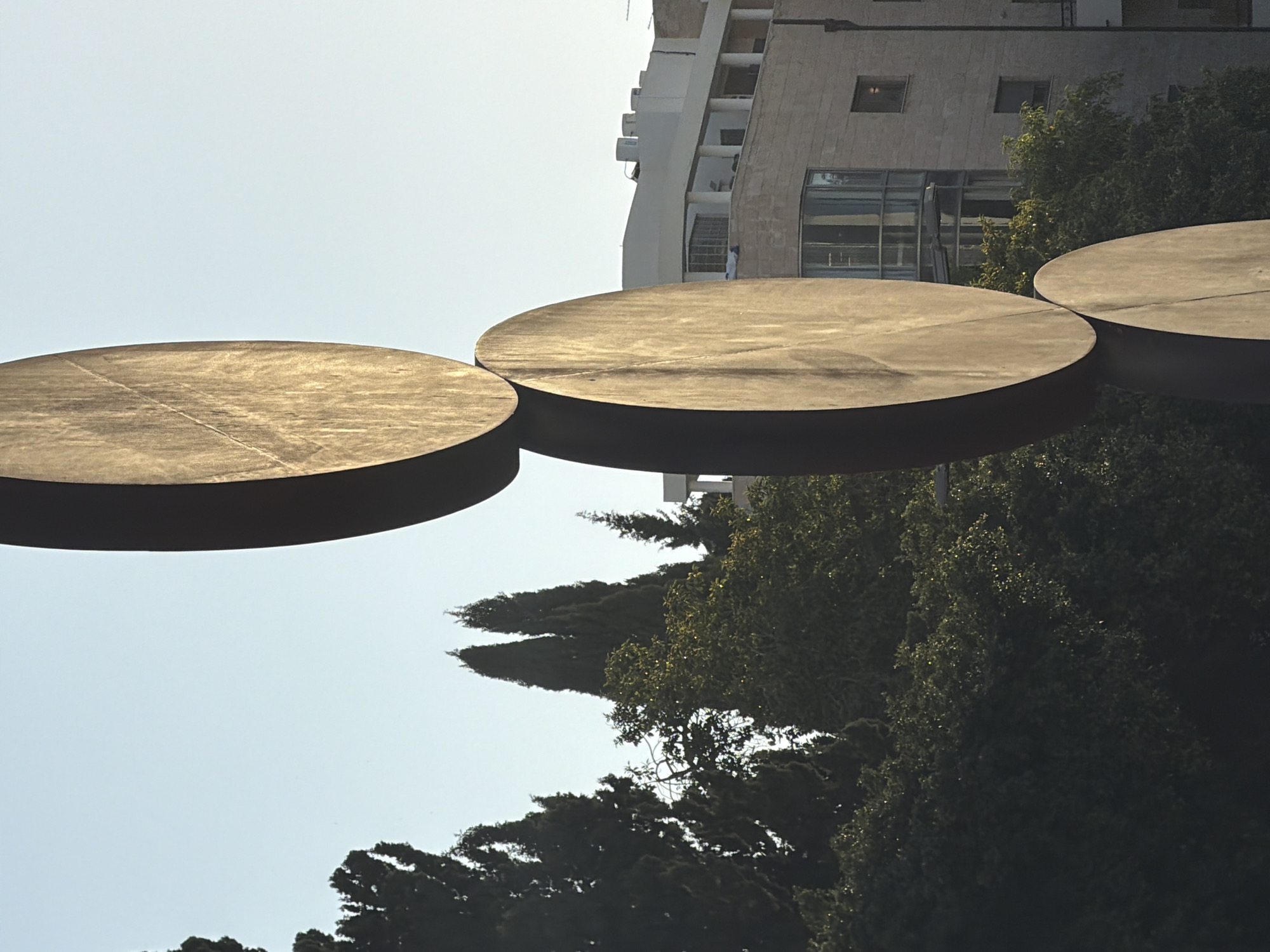 The image captures a low-angle view of "Turning Point," a kinetic sculpture by Israeli artist Beverly Pepper, located in Tel Aviv, Israel. The artwork consists of three large, circular disks suspended in the air. The disks have a weathered, golden-brown metallic surface.
Behind the sculpture, there is a clear, light blue sky. A building is visible in the background with light colored facades, windows and balconies. The trees are densely packed together, creating a dark green backdrop. The image is rotated, placing the ground on the right side of the frame.