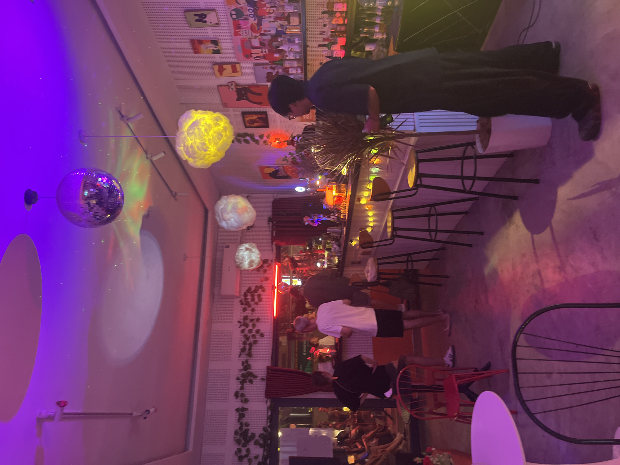 The image depicts the interior of a bar in Da Nang, Vietnam, illuminated by a combination of purple, pink, and red lights. A disco ball hangs from the ceiling, reflecting light across the space. Several patrons are visible, some seated at tables and others standing near the bar. The bar itself is stocked with bottles, and decorative lights adorn its front. In the foreground, a person in dark clothing stands facing away from the camera, holding a bottle. The walls are decorated with a variety of framed artwork and a vine-like garland. Chairs and small tables are arranged throughout the bar area.