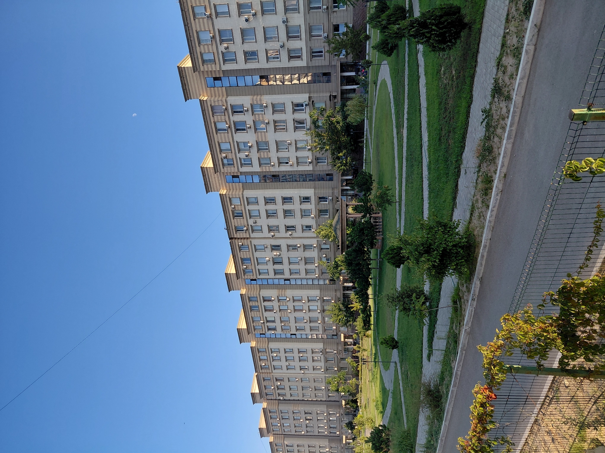 The image shows a modern, multi-story residential building with a light-colored facade and decorative beige trim. The building features numerous windows and air conditioning units. In front of the building, there is a well-maintained park with green lawns, curved pathways, and scattered trees and shrubs. A gazebo is visible in the park. A paved road runs along the edge of the park, separated by a fence and vegetation. The sky is clear and blue, with a visible crescent moon. This scene is typical of the residential areas in Aktau, Kazakhstan, a coastal city known for its modern architecture and emphasis on green spaces within urban environments.
