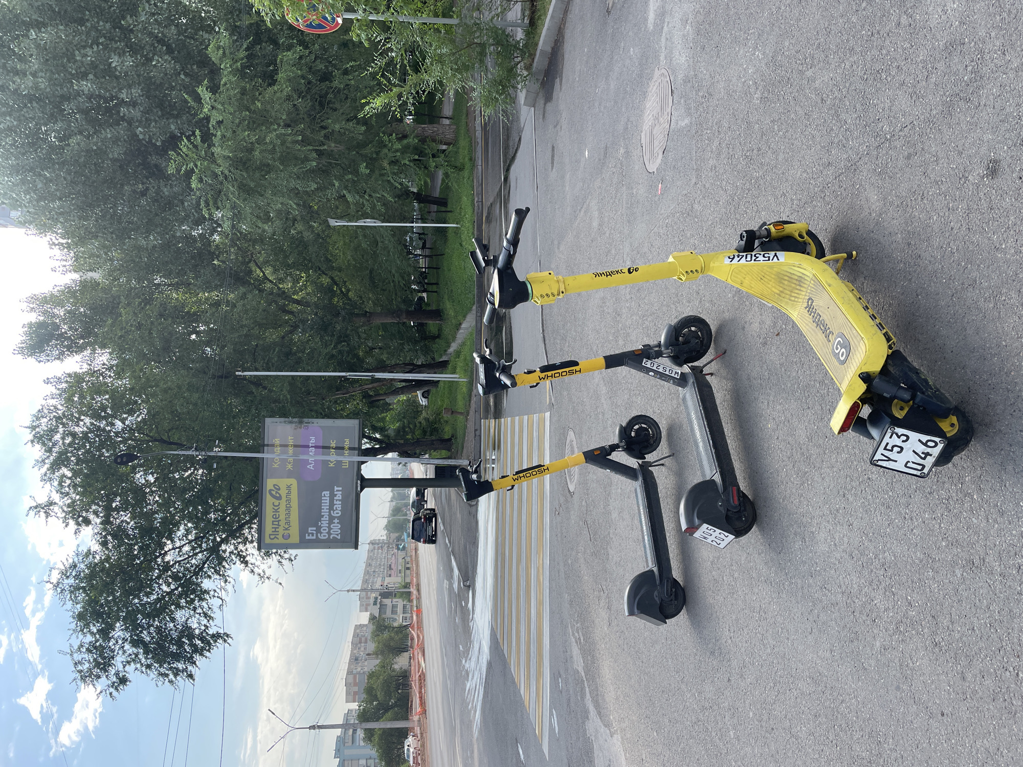 In Almaty, Kazakhstan, on a cloudy day, two yellow and black electric scooters are parked on an asphalt street next to a pedestrian crossing. The scooter in the foreground has the license plate Y53 046 and the word "Whoosh" printed on its side. The other scooter is slightly behind it and also features the "Whoosh" branding. A large billboard for Yandex Go is visible in the background, advertising services in various locations. Trees line the street, and buildings are visible in the distance. The street appears wet, possibly from recent rain. A few cars are driving on the road, and there is construction fencing on the side of the street. The overall scene depicts a typical urban environment with readily available transportation options.
